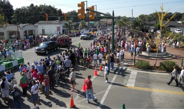 Crowd of people waiting at a street corner, with some crossing the street and others standing near a traffic light and orange cones, in a busy urban area with shops and a small park.