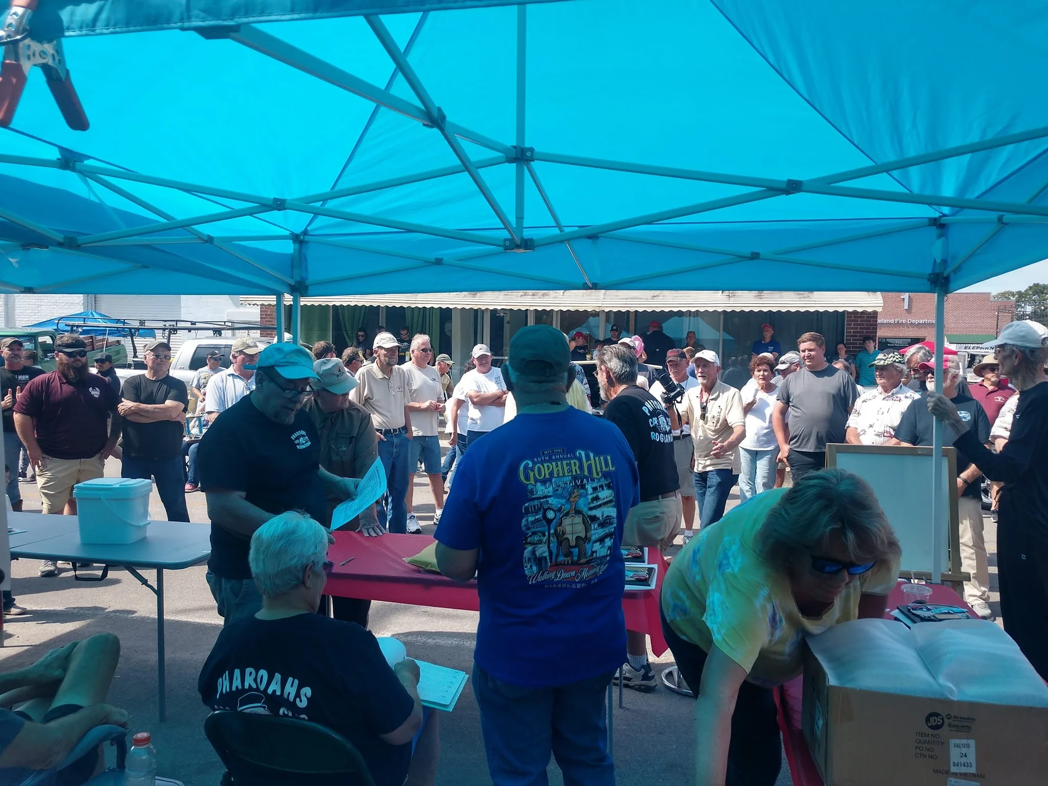 A crowd of people gathered outdoors under a blue canopy tent, some are standing and others are sitting at tables, during a festival or community event.