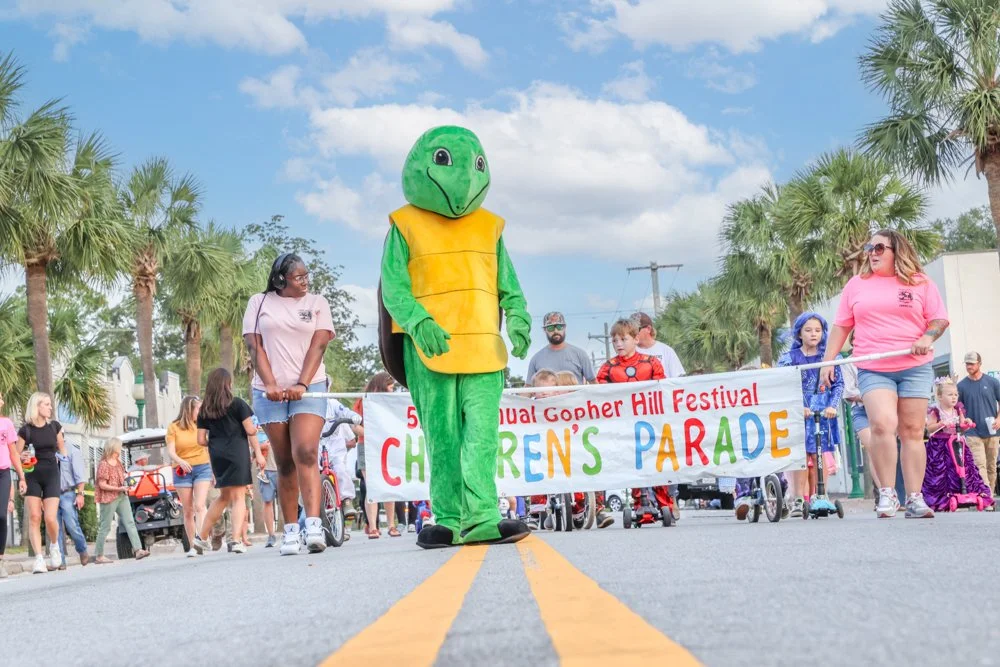 Person dressed in a green turtle costume marching in a children's parade during Gopher Hill Festival, with people and children walking behind a banner.