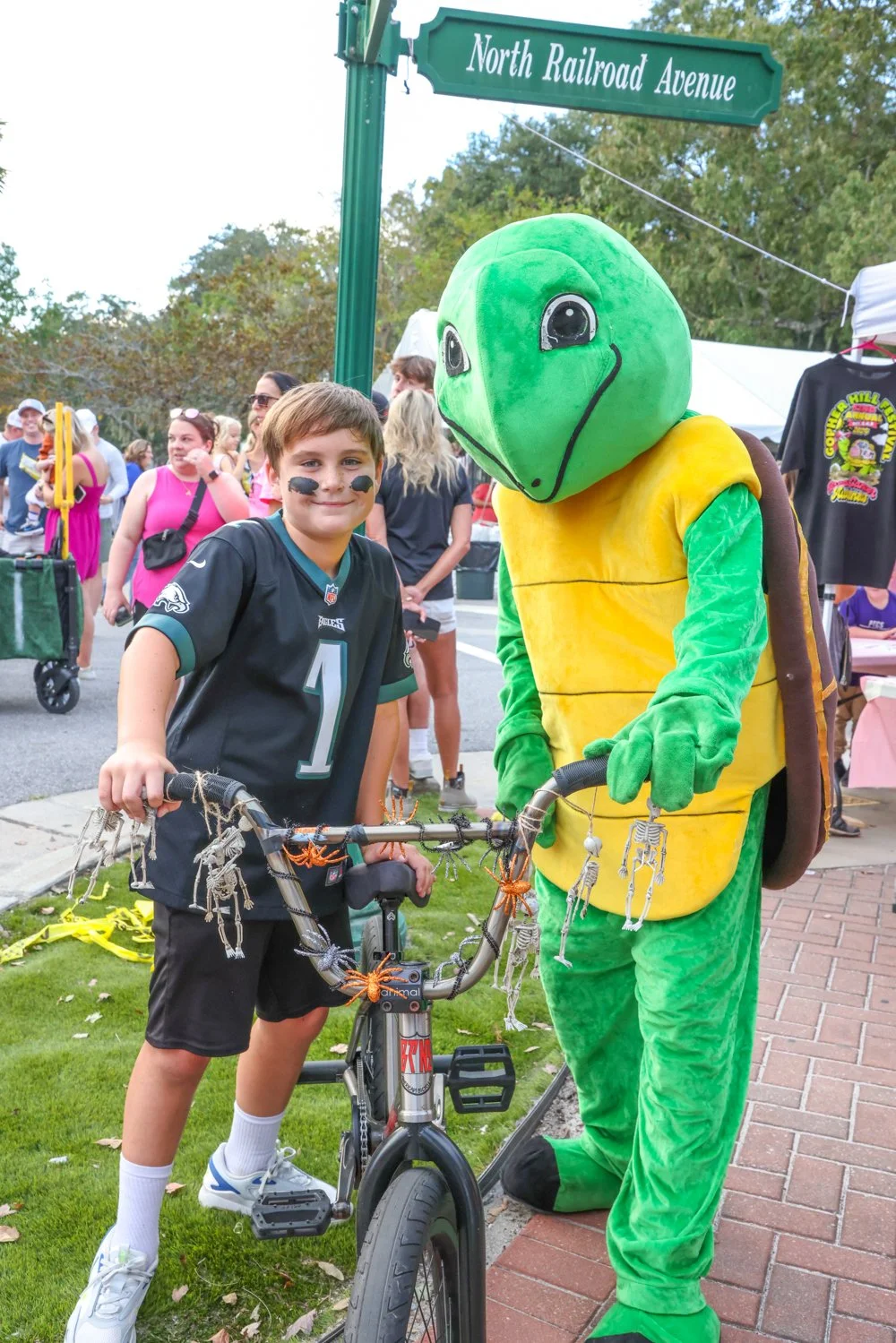 A young boy with face paint and a black jersey is standing next to a person in a turtle costume, holding a decorated bike at an outdoor event. People are gathered in the background and a street sign reads 'North Railroad Avenue.'