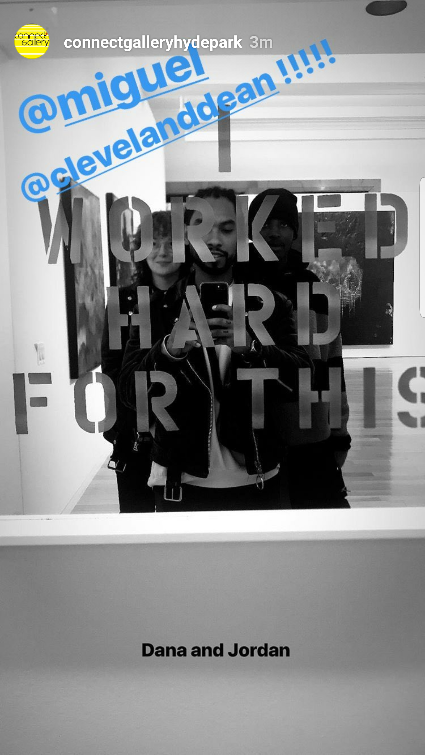 Black and white photo of three young adults, two men and a woman, taking a mirror selfie in an art gallery or similar setting. The mirror has a cutout design with the words "WORKED HARD FOR THIS." The woman is holding a smartphone. The printed captio