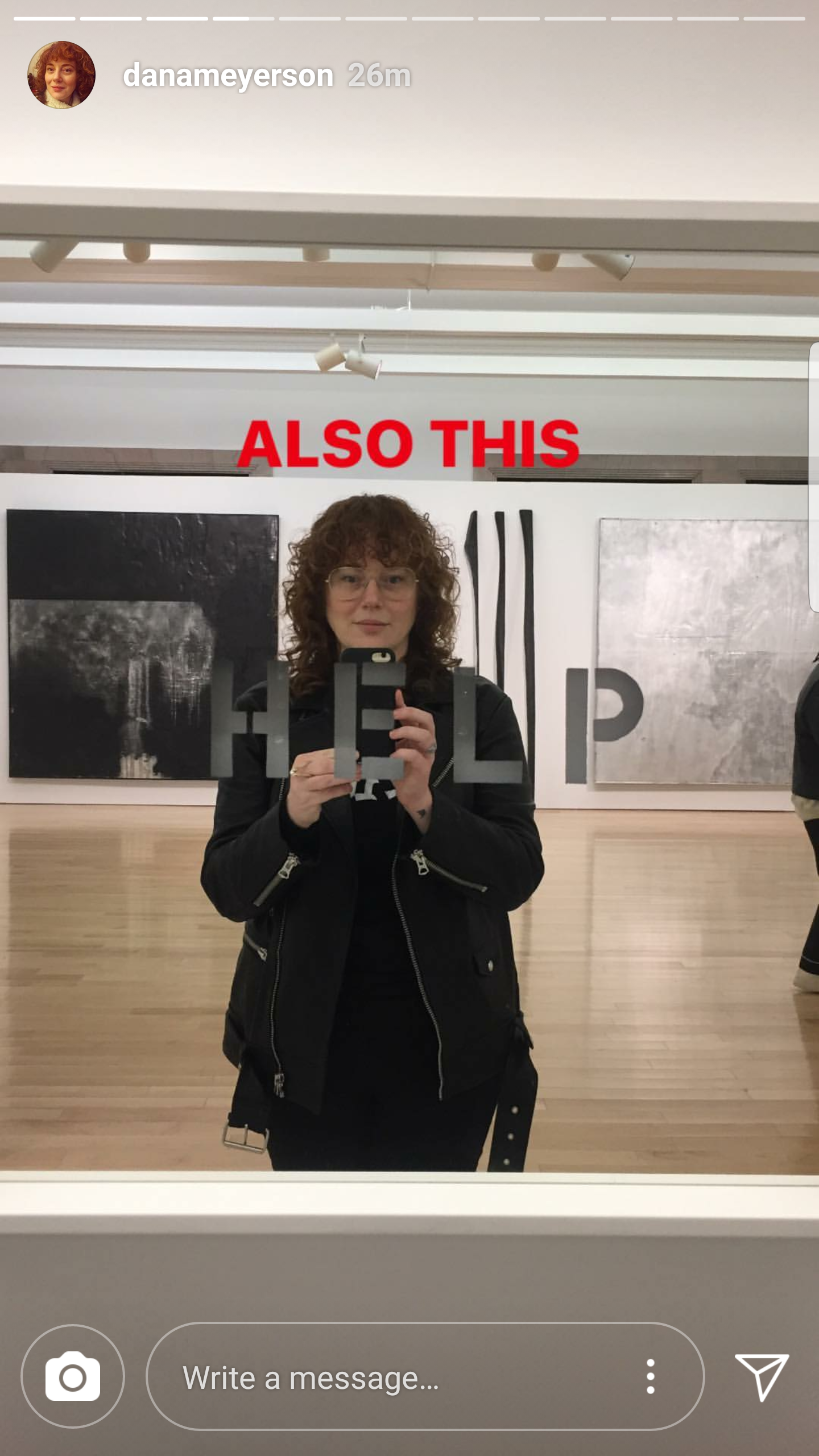 A woman with curly red hair and glasses taking a mirror selfie in an art gallery, holding a phone with the letters 'HELP' visible on the mirror, with artwork in the background and the text 'ALSO THIS' in red above her head.