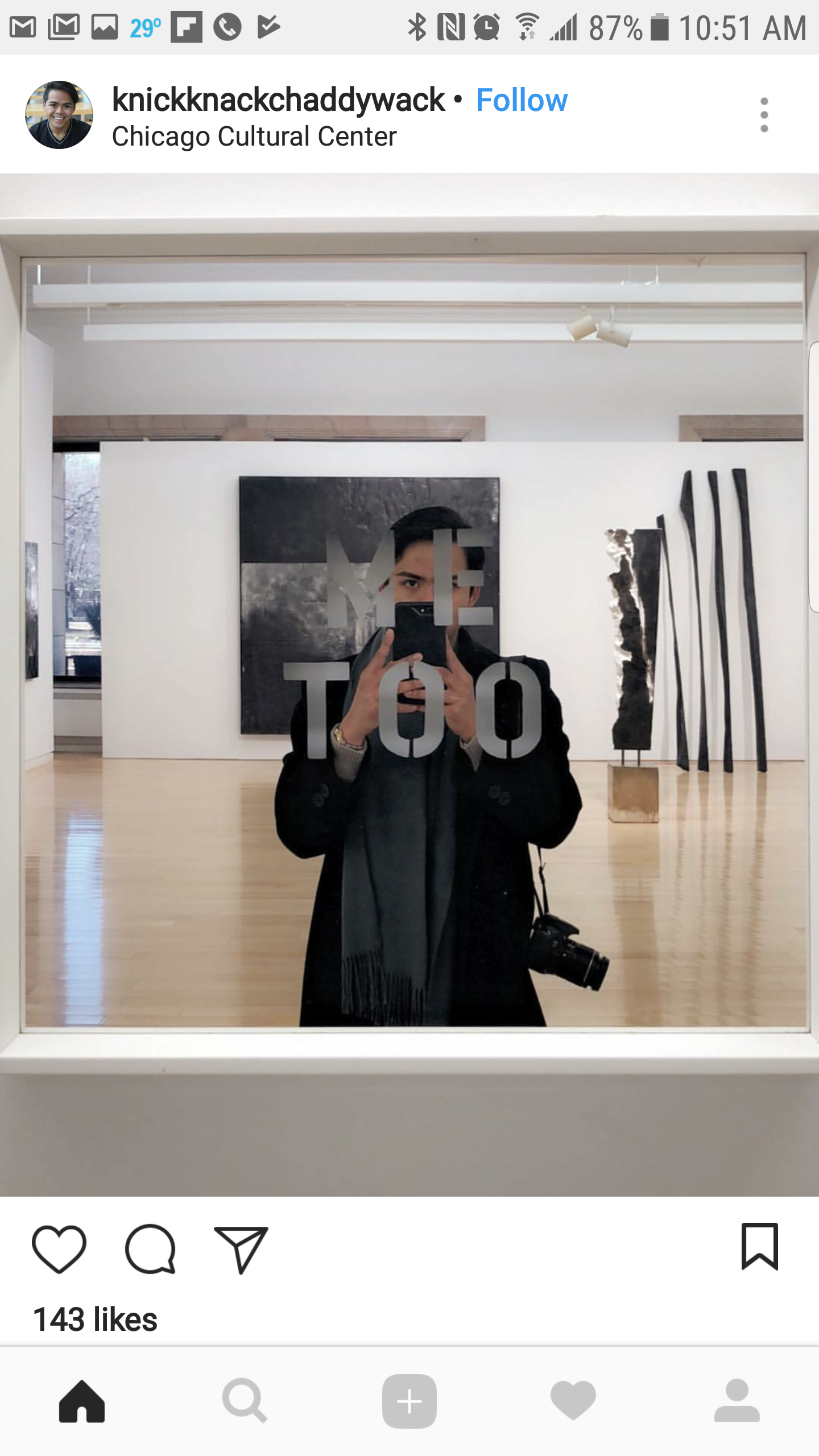A person taking a selfie in an art gallery mirror that has the words "THE ART ROOM" on it. The person is dressed in dark clothing and holding a camera with some artwork visible in the background.