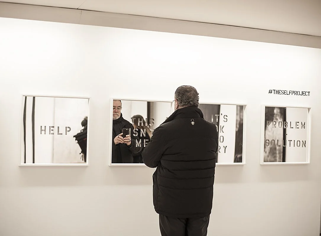 A man with glasses and a black jacket looking at framed art pieces on a gallery wall, with his reflection visible in the middle mirror.