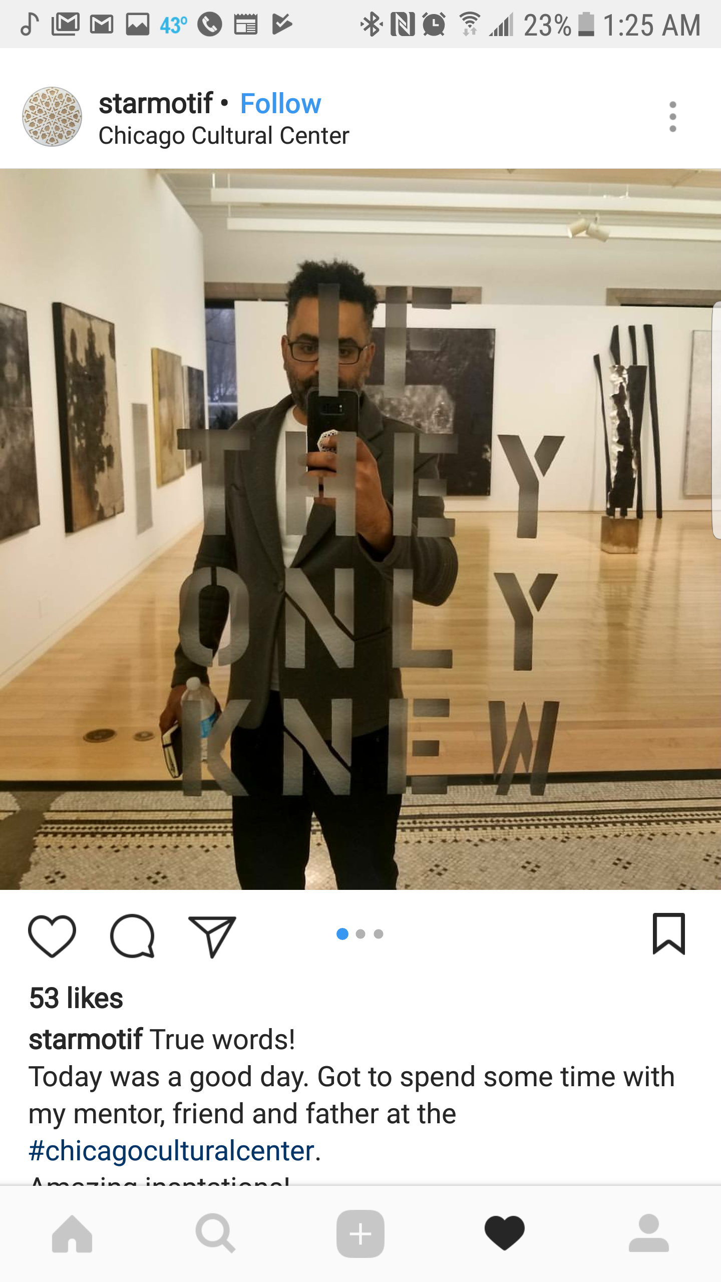 Person taking a selfie in an art gallery, reflected in a mirror with black acrylic decals spelling "THE ONLY NEW." The gallery has white walls, abstract art on display, and a wooden floor.