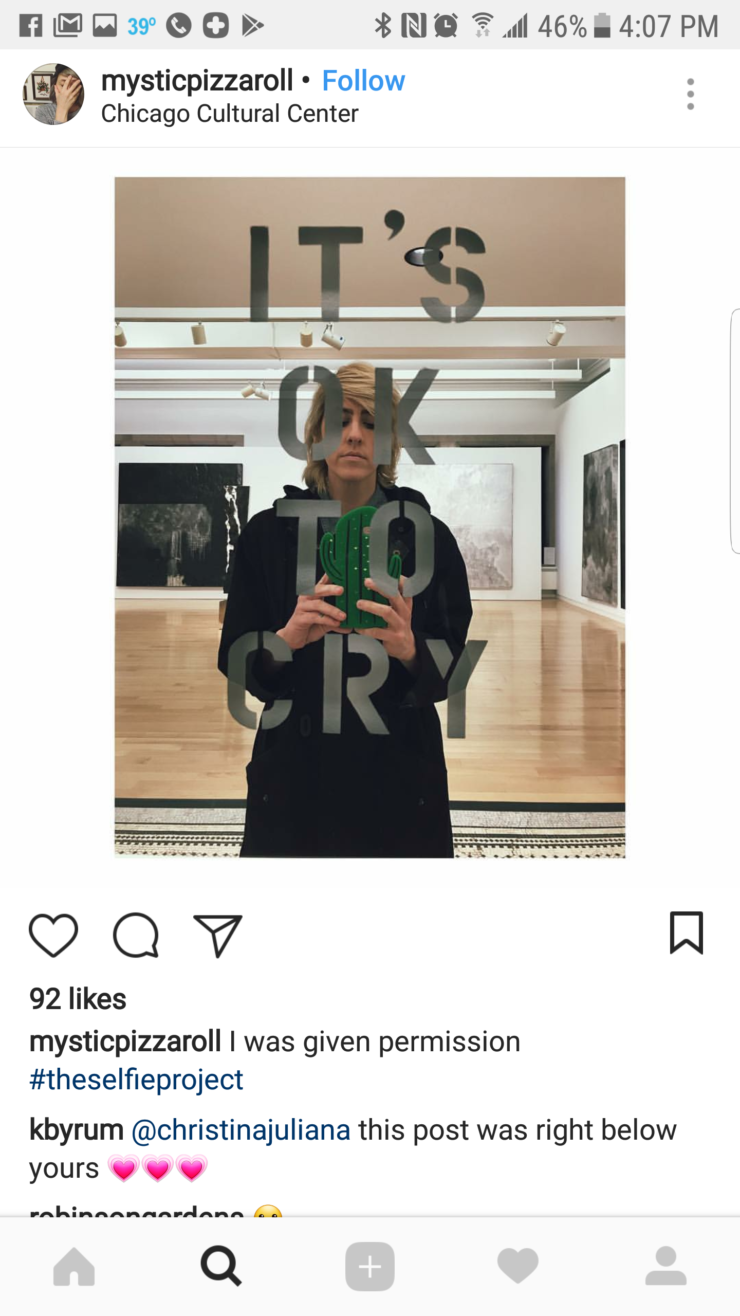 A woman taking a mirror selfie in an art gallery at the Chicago Cultural Center. The mirror has the words 'IT'S OK TO CRY' written on it, partly obscuring her face. The gallery features artwork on the walls and a wooden floor.