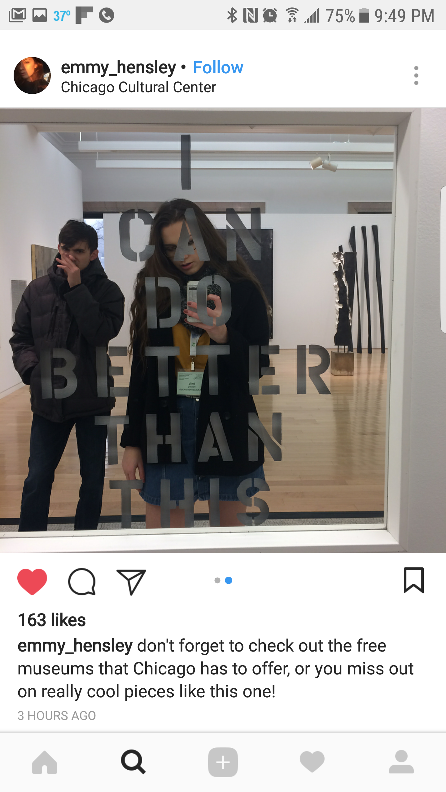People taking a selfie in front of a mirrored window with an inscription, inside a museum gallery with wooden floors and abstract sculptures in the background.