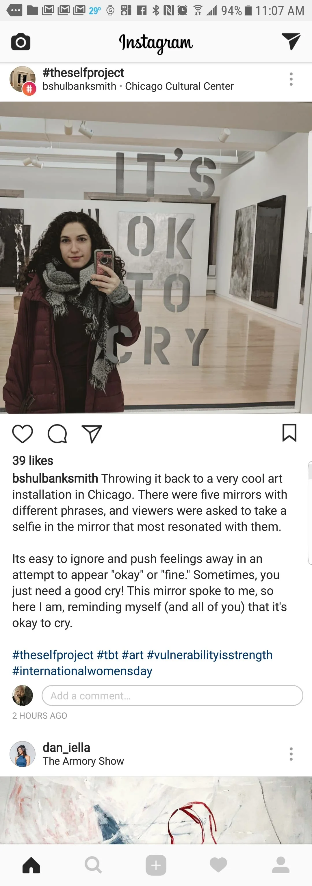 A woman taking a selfie in an art gallery with the words 'IT'S OK TO CRY' displayed on the wall behind her.