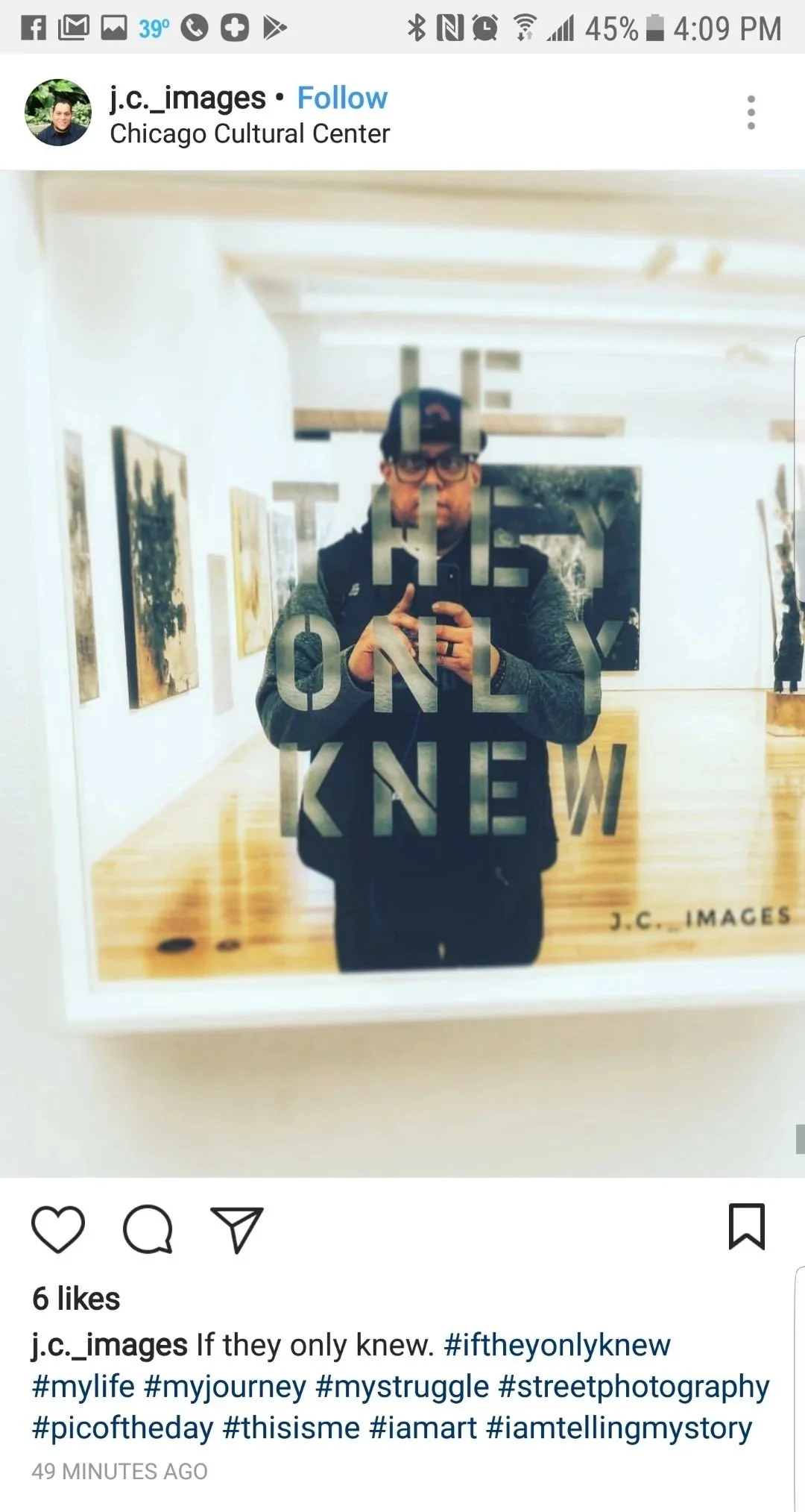 A person taking a mirror selfie in an art gallery with paintings on the wall behind them, through a glass that has words etched on it, including 'THE ONLY NEW'.