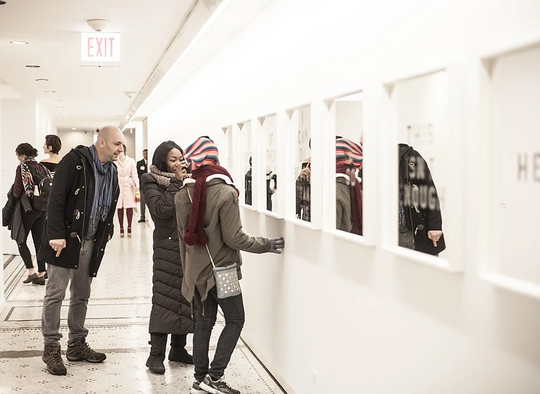 People observing small artworks through window-like openings in a white gallery wall.