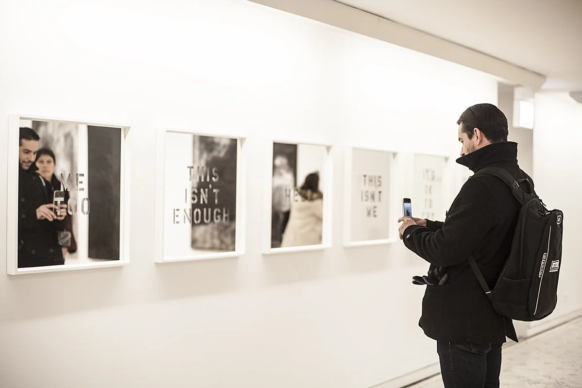 A man in a black coat and backpack taking a photo with his phone in an art gallery. The gallery wall displays frames with images and text, some reads "THIS ISN'T ENOUGH."