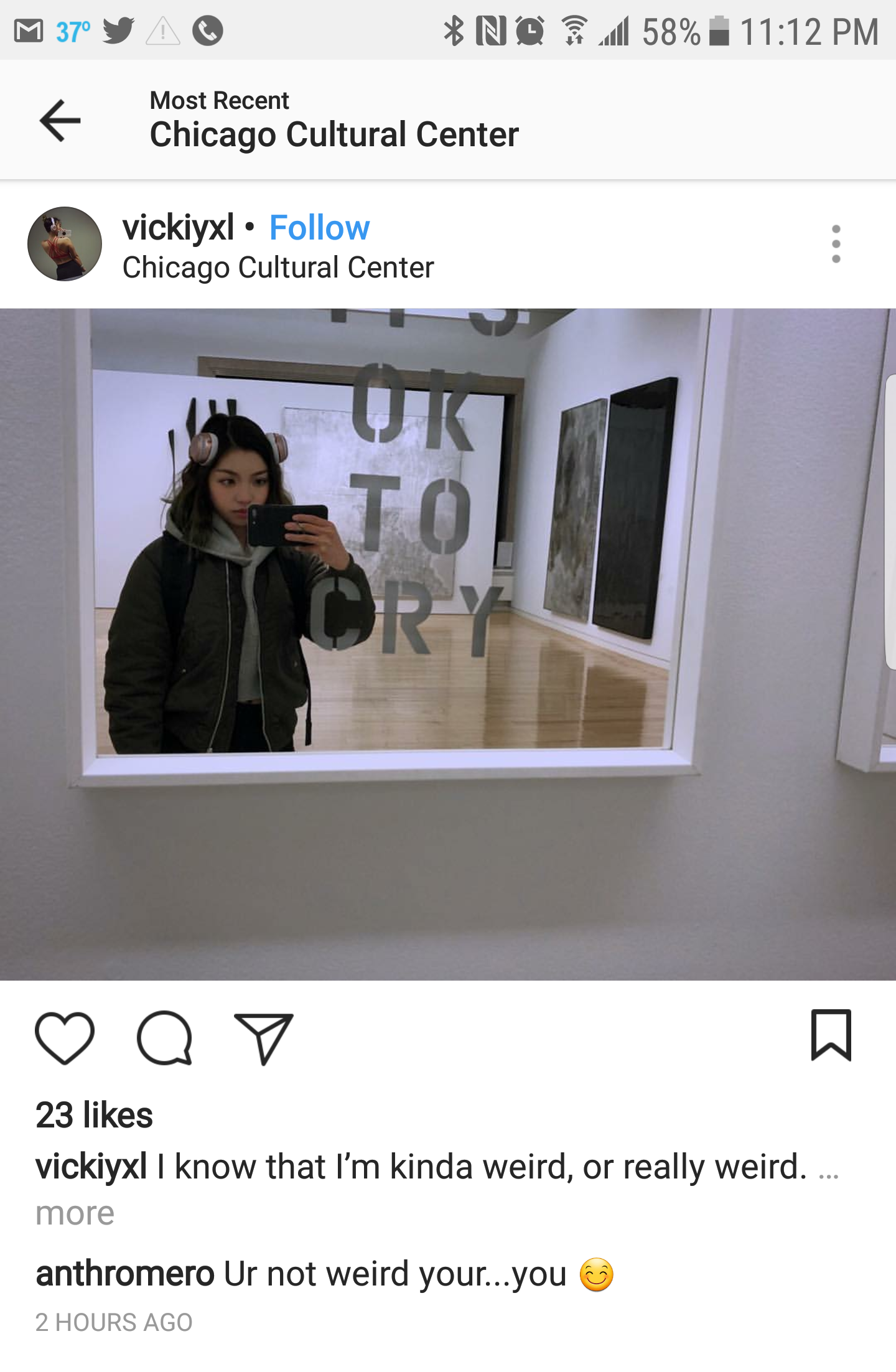A person taking a selfie in front of a mirror at the Chicago Cultural Center. The mirror has the words 'LOOK TOY' written on it vertically. The person is wearing headphones, a jacket, and a backpack.