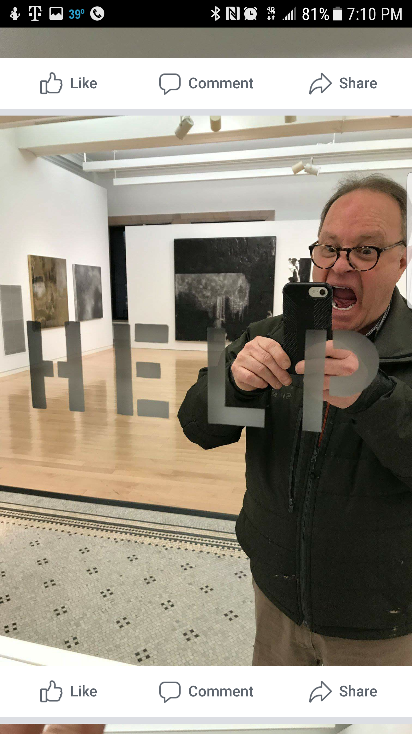 Man taking a photo in art gallery with artworks on the walls, reflected in a glass door with a sign.