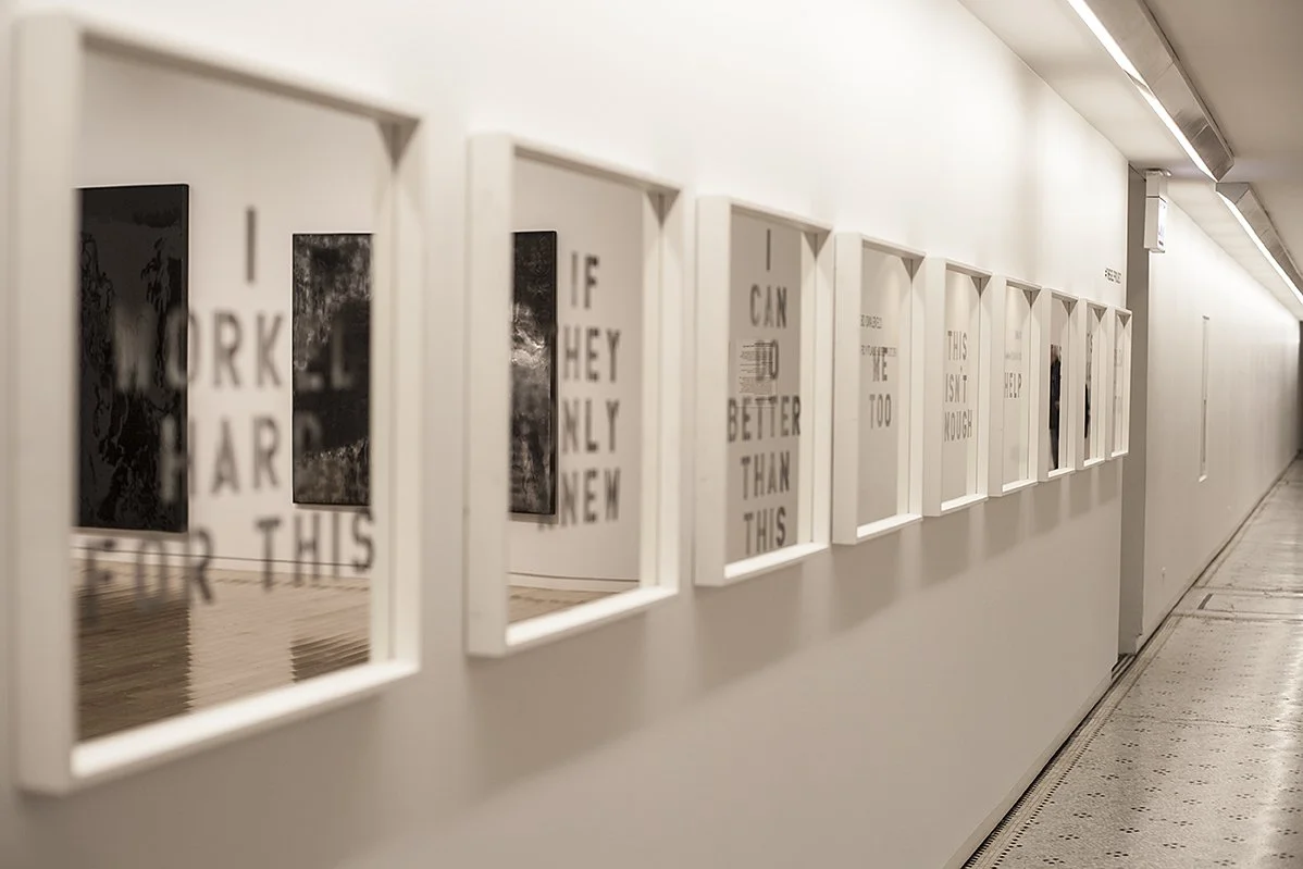 Gallery wall with framed text artworks in white frames along a hallway with a white wall and ceiling lights.