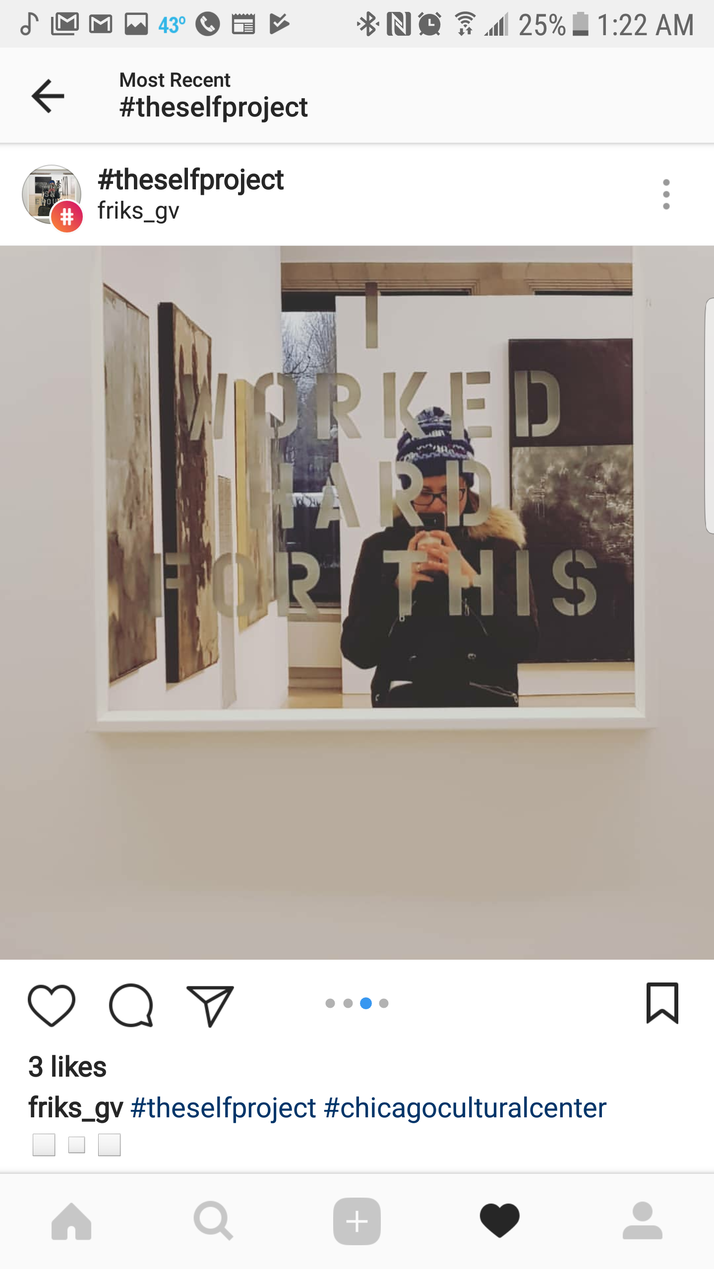 A person in a winter hat with a pom-pom and glasses taking a selfie in a mirror at an art gallery. The mirror has a quote that reads, 'WORKED HARD FOR THIS.' Behind the person, there are abstract paintings on the wall and a window showing trees outsi