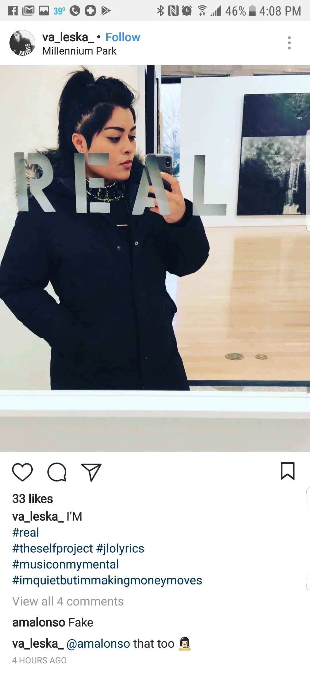 A woman taking a mirror selfie at Millennium Park, wearing a black jacket with the word 'REAL' reflected in the mirror, and dark hair styled in a high ponytail. The background shows a wooden wall and framed photographs.