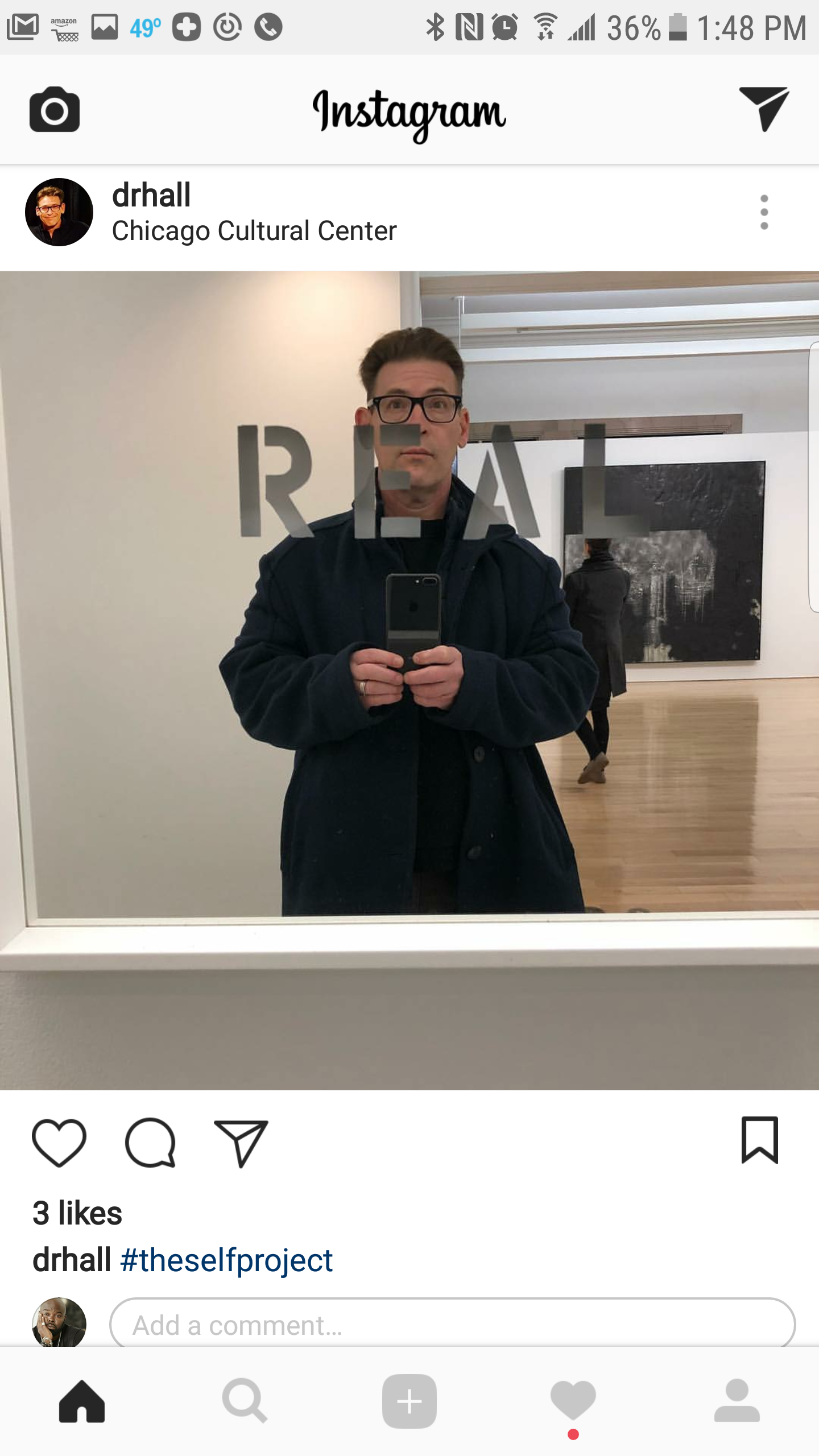 A man taking a selfie in front of a mirror at the Chicago Cultural Center. The mirror has the word 'REAL' partially visible. The man is wearing glasses and a dark coat, and another person is seen walking in the background.