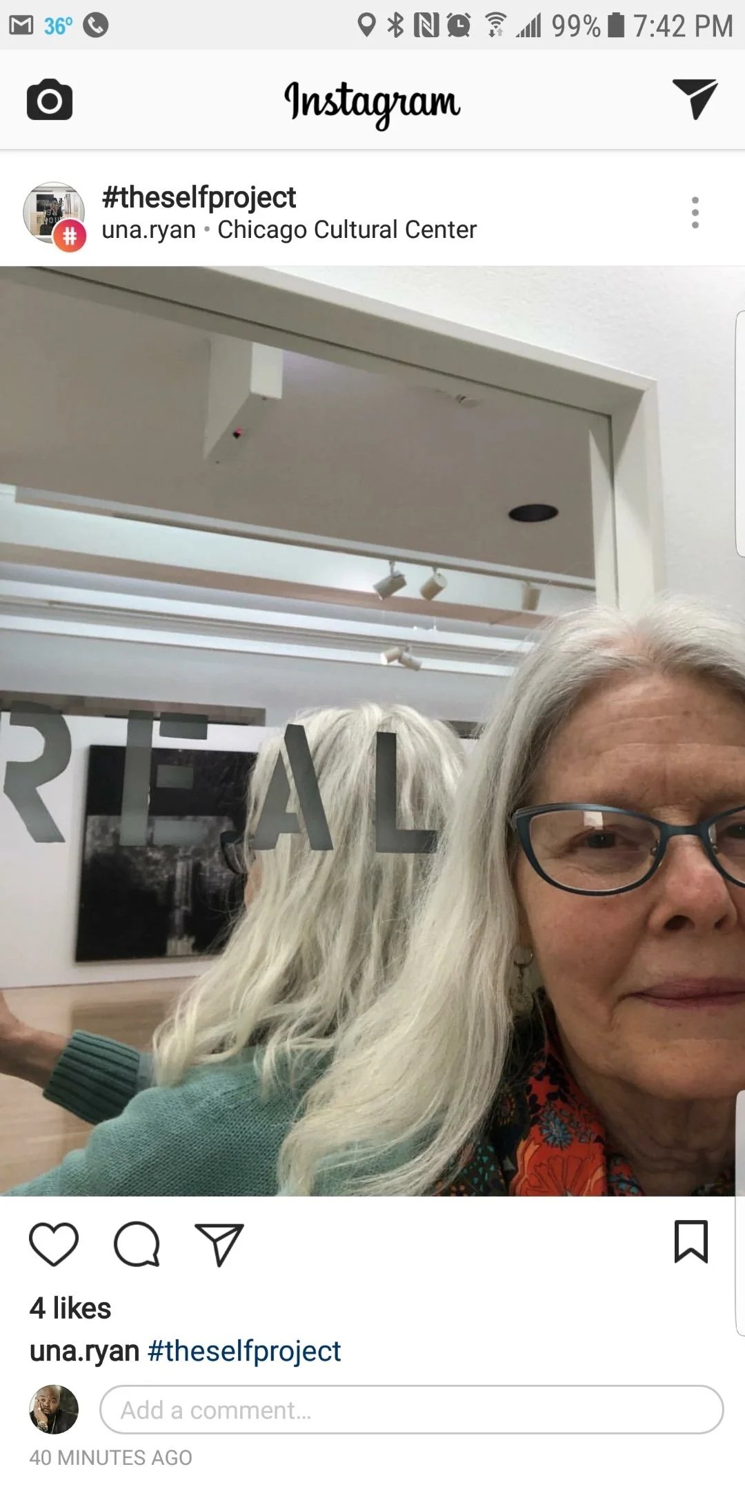 A woman with gray hair and glasses taking a selfie in front of a glass door with the word 'TRIAL' on it, inside an art gallery or cultural center.