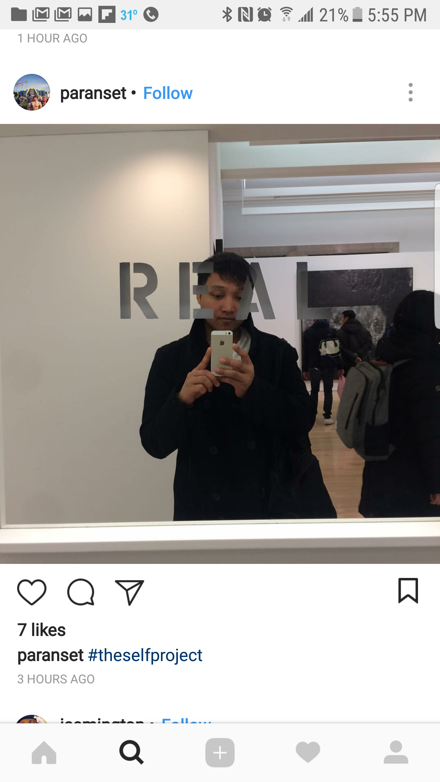 A person taking a selfie in a mirror at an indoor location with several people in the background. The reflection shows a sign with the partial word 'REAL' behind him.