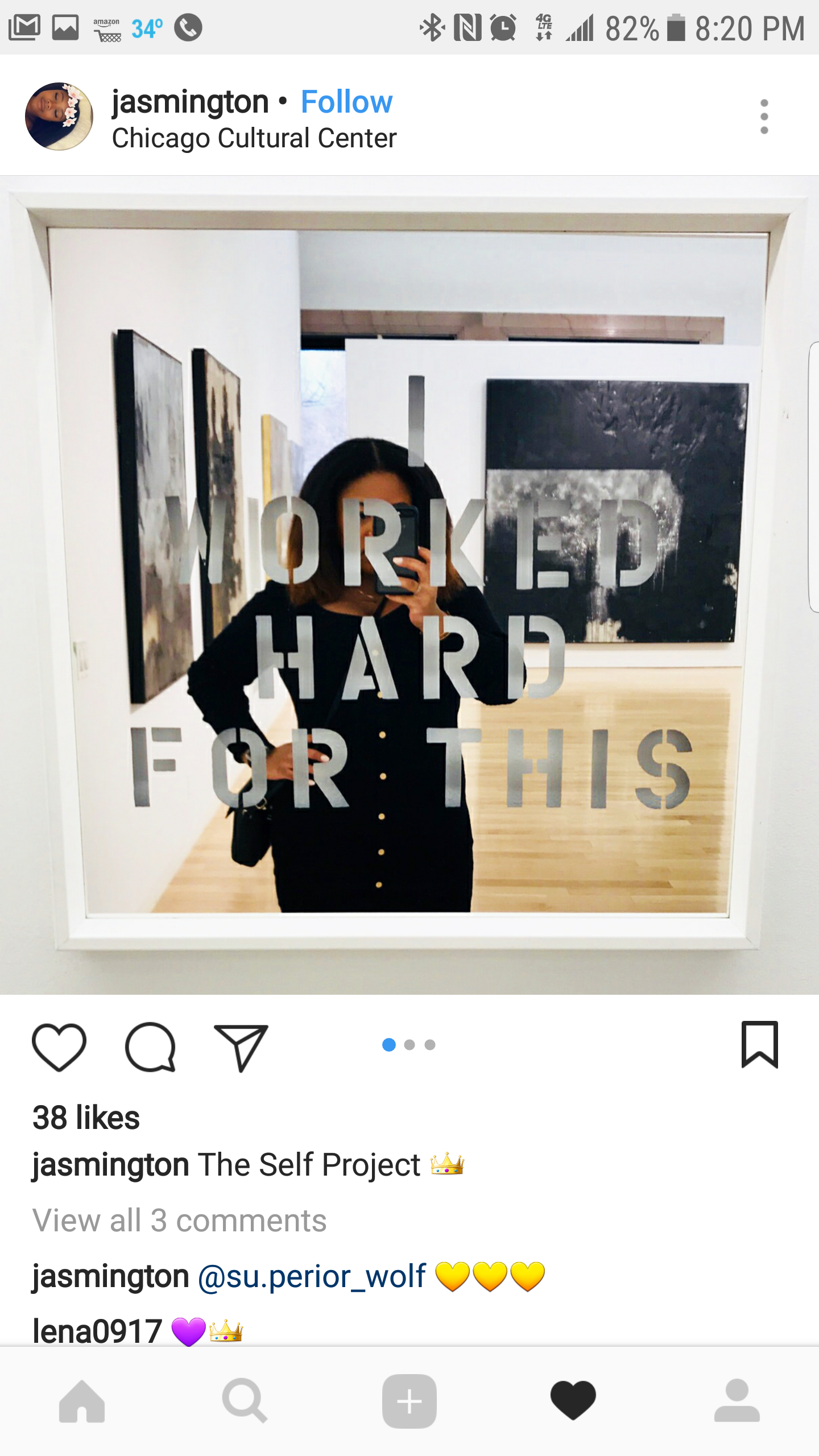 A woman taking a mirror selfie in an art gallery, with painted artworks visible on the walls behind her. The mirror has the words "WORKED HARD FOR THIS" written on it.