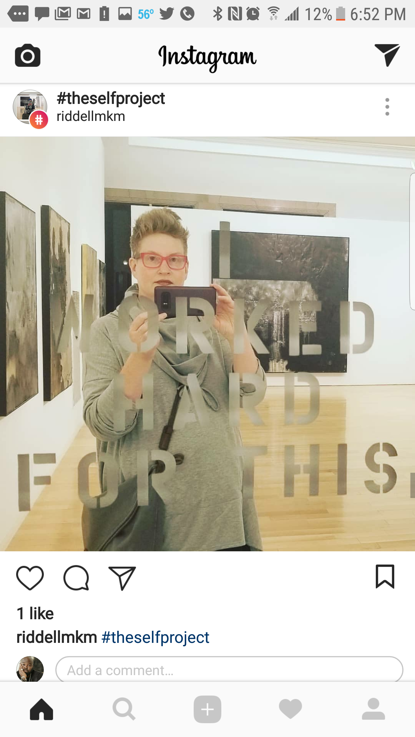 A woman taking a mirror selfie in an art gallery, wearing glasses and a gray jacket, with artworks displayed on the wall behind her.
