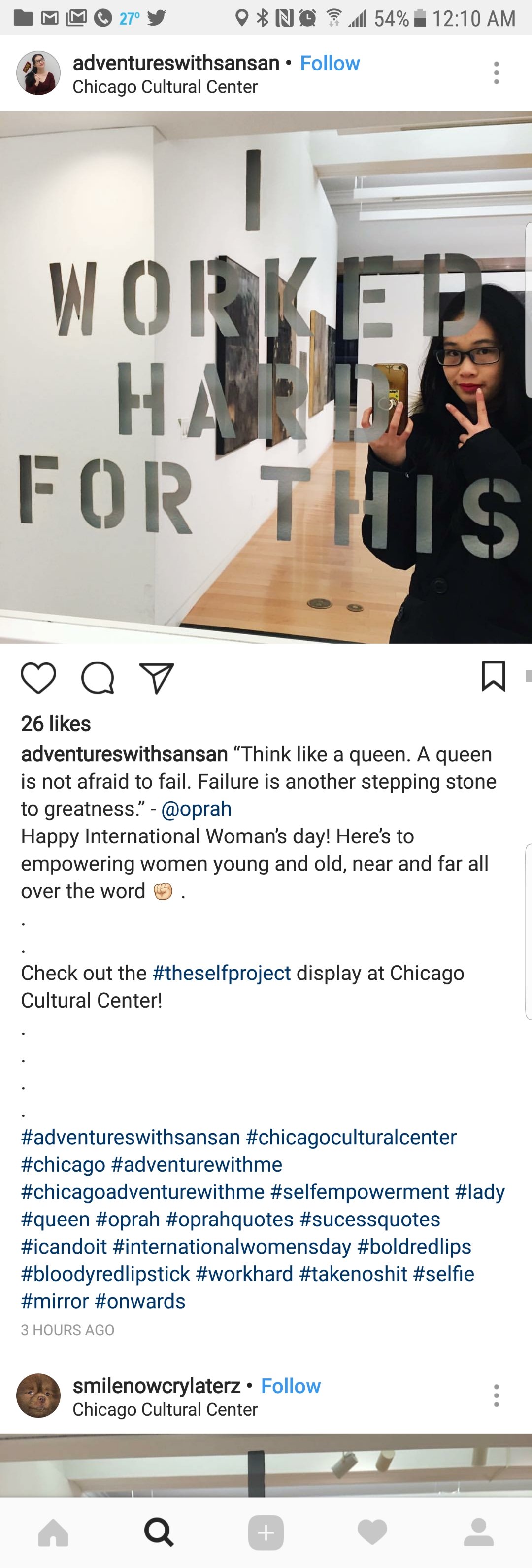 A mirror with cut-out words reading "WORKED HARD FOR THIS." A woman with glasses and dark hair is taking a selfie in front of the mirror, holding her phone with her right hand and making a peace sign with her left hand. The background shows paintings