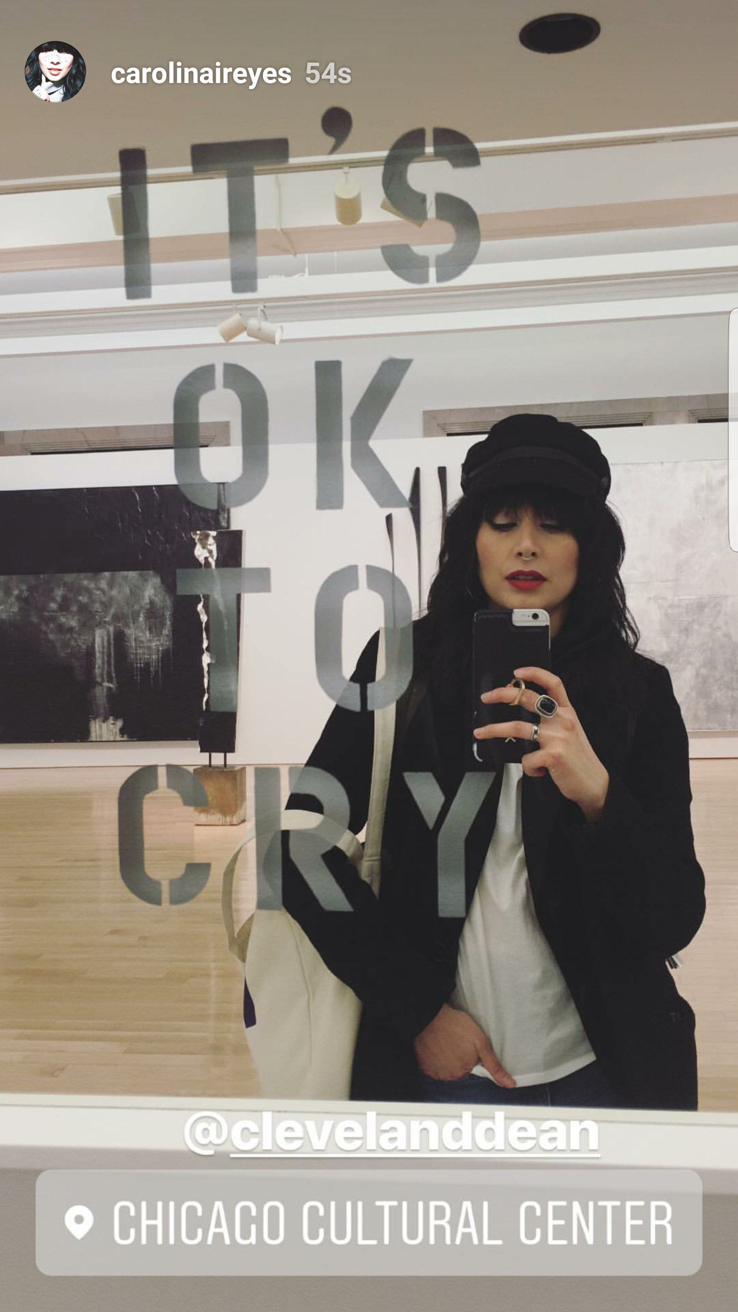 A woman taking a selfie in a mirror at the Chicago Cultural Center, wearing dark clothing and a black hat, with art galleries visible in the background.
