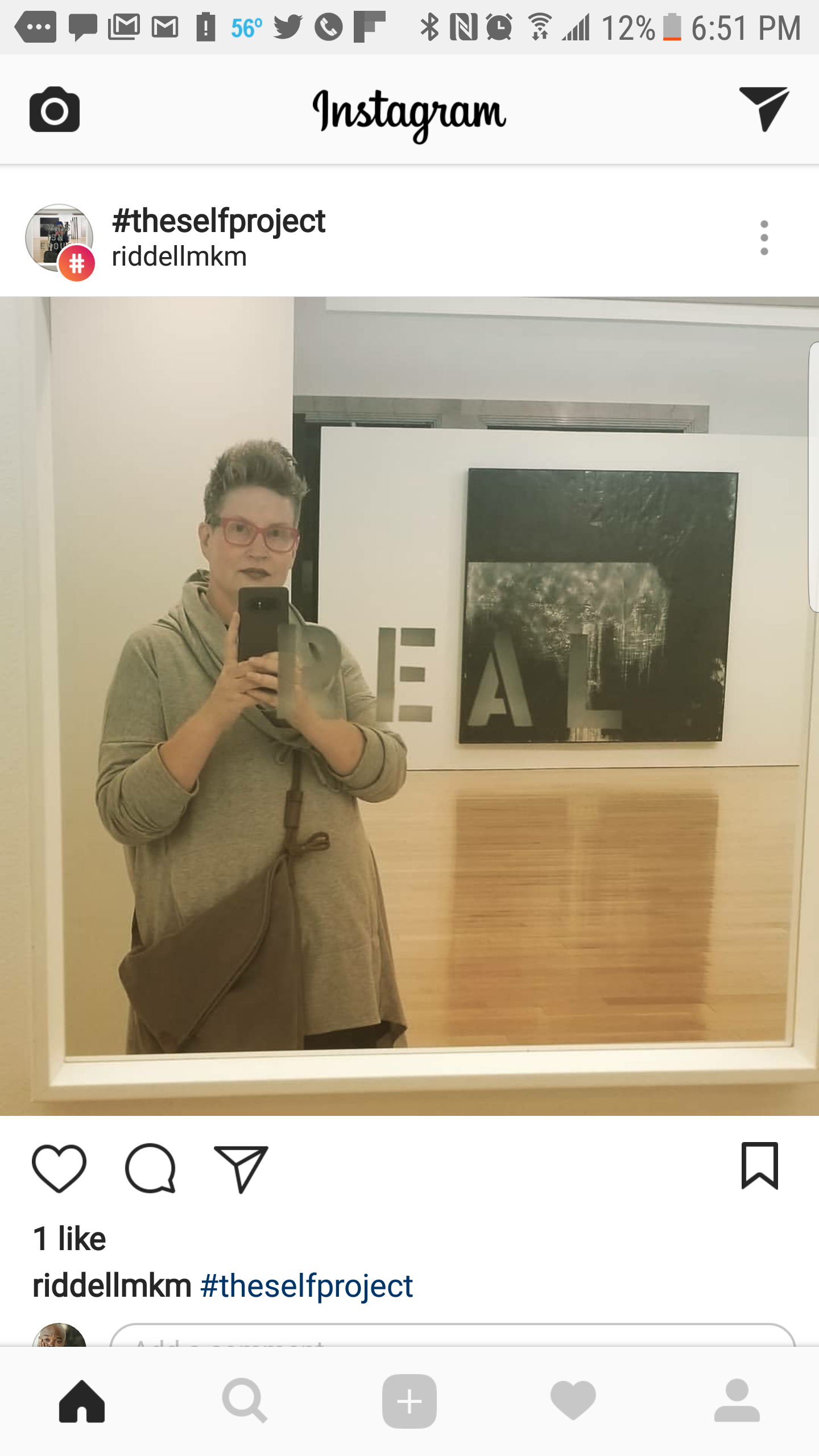 A woman taking a selfie in a mirror at an art exhibit, standing in front of a black and white artwork with the letters turning by the reflection, wearing glasses and a gray hoodie.