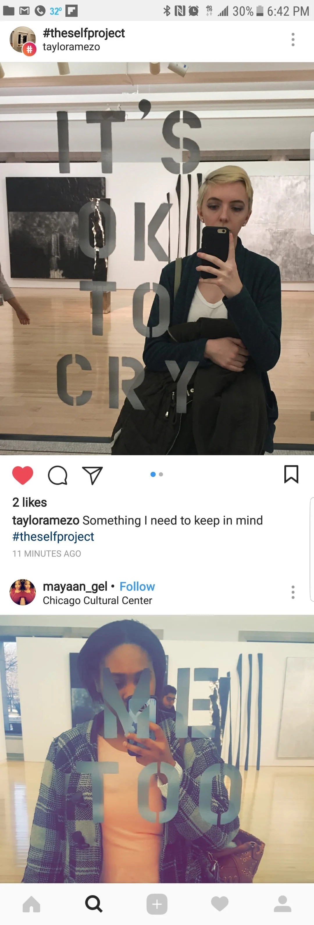 Two women taking mirror selfies at an art gallery, with large abstract paintings in the background. The mirror has the words 'IT'S OKAY' written on it.