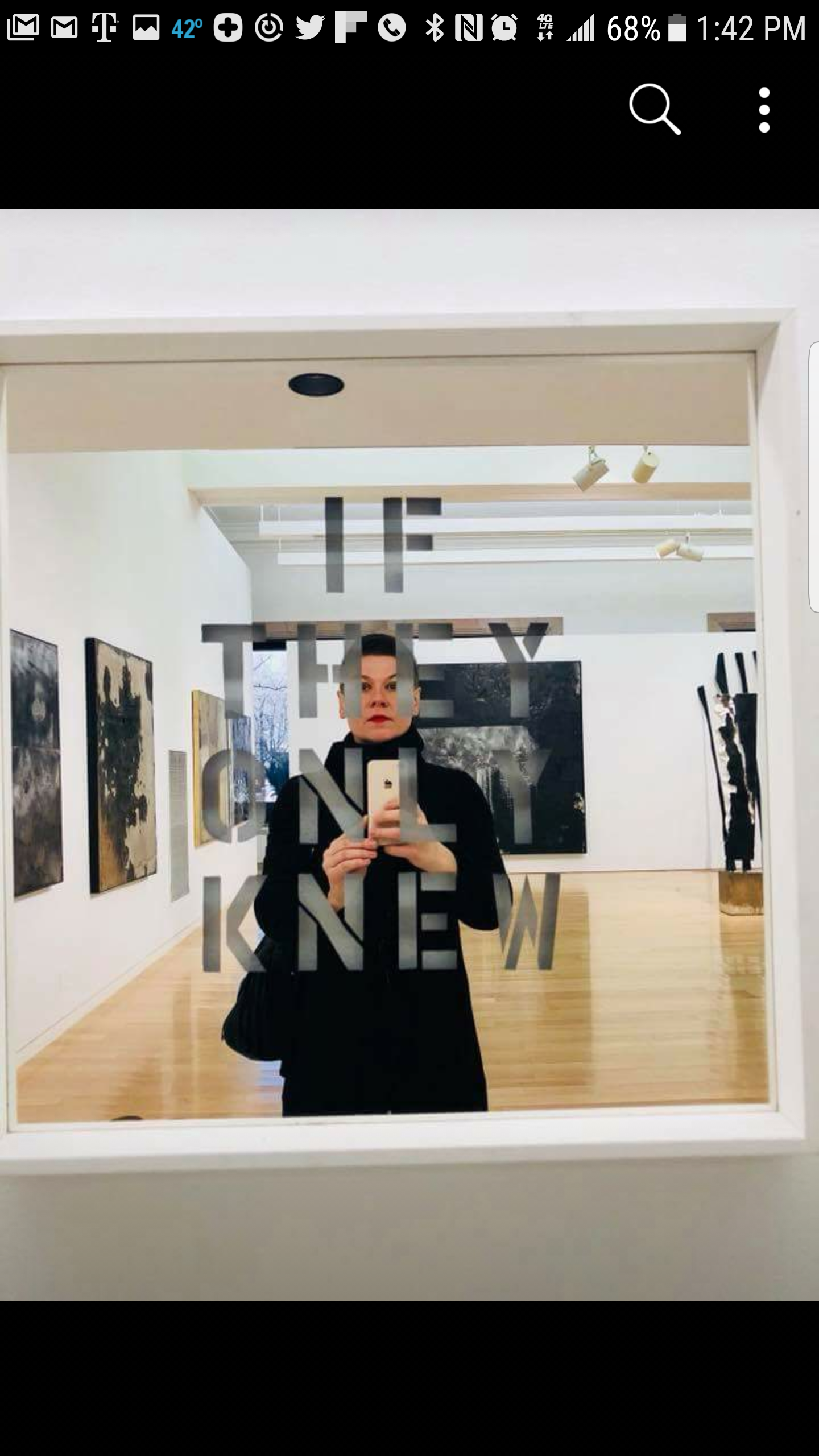 Person taking a mirror selfie in an art gallery, wearing black, with the words 'THE EMPTY GALLERY' reflected on the mirror, surrounded by artworks and sculptures.
