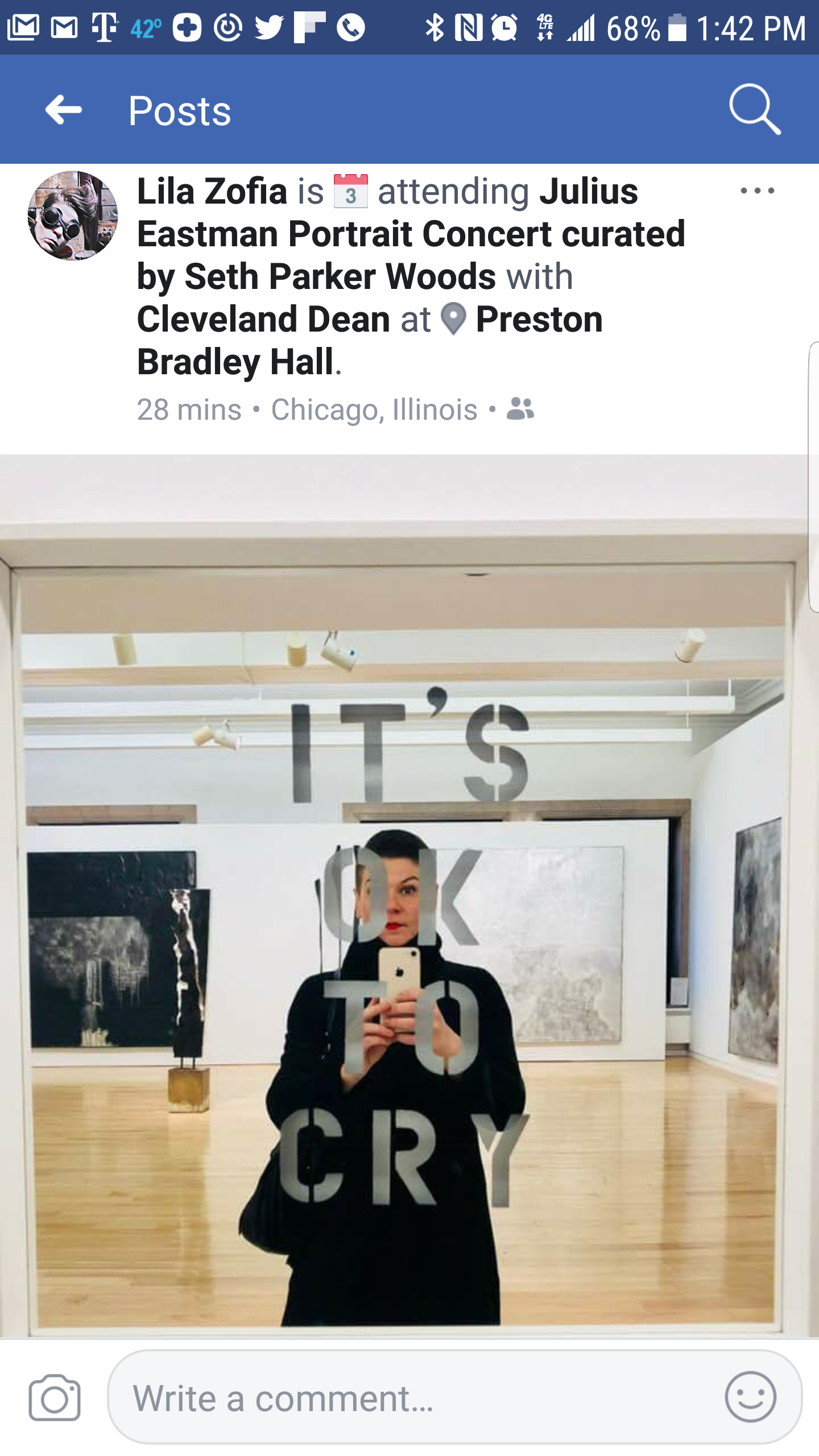 A woman taking a selfie in front of a mirror with the phrase 'IT'S OK TO CRY' written on it, in an art gallery with wooden floors and abstract paintings on the walls.