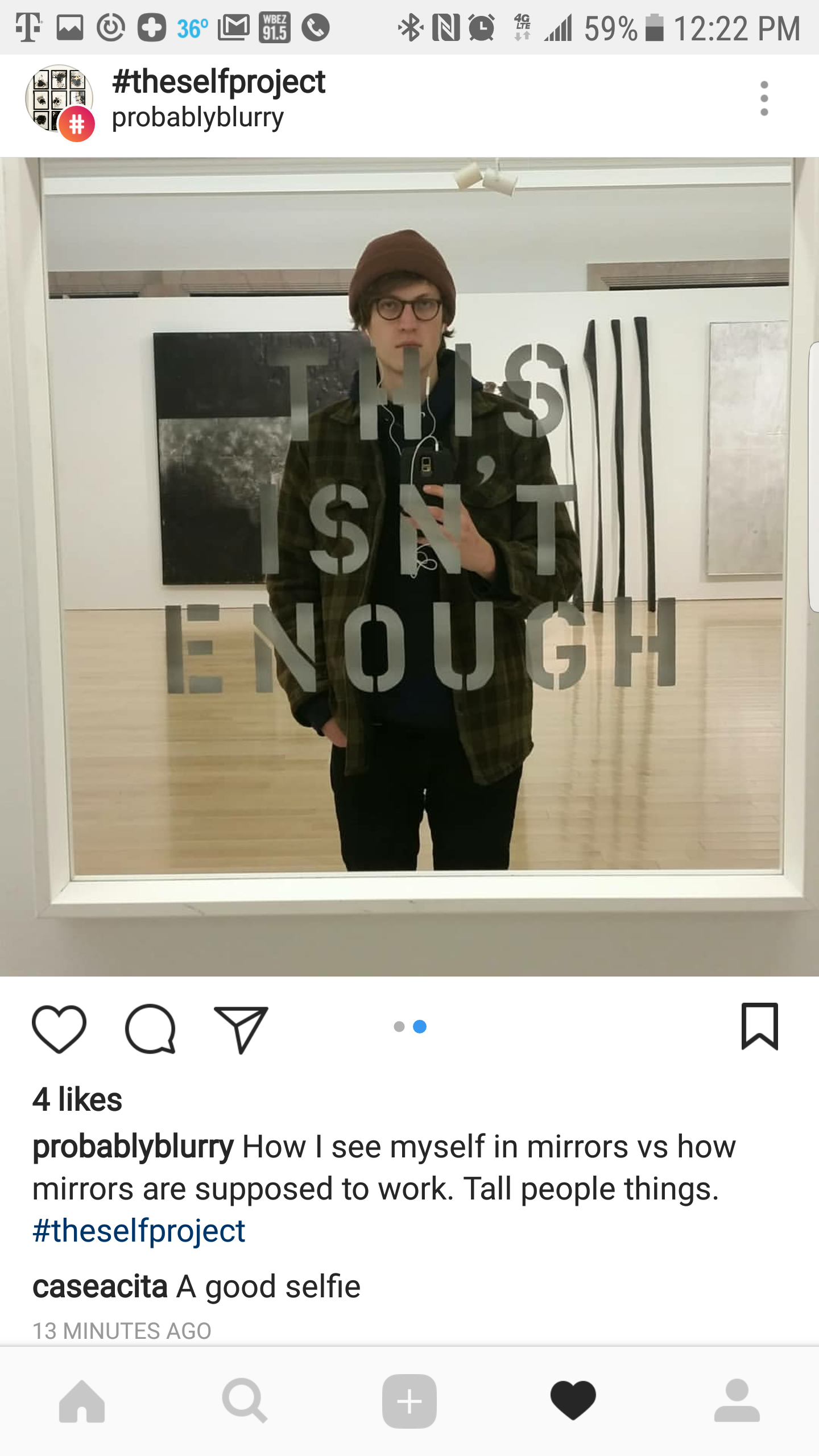 A person taking a mirror selfie in an art gallery, with a sign on the mirror that reads 'THIS ISN'T ENOUGH'. The person is wearing glasses, a brown beanie, a dark hoodie, and a plaid jacket.