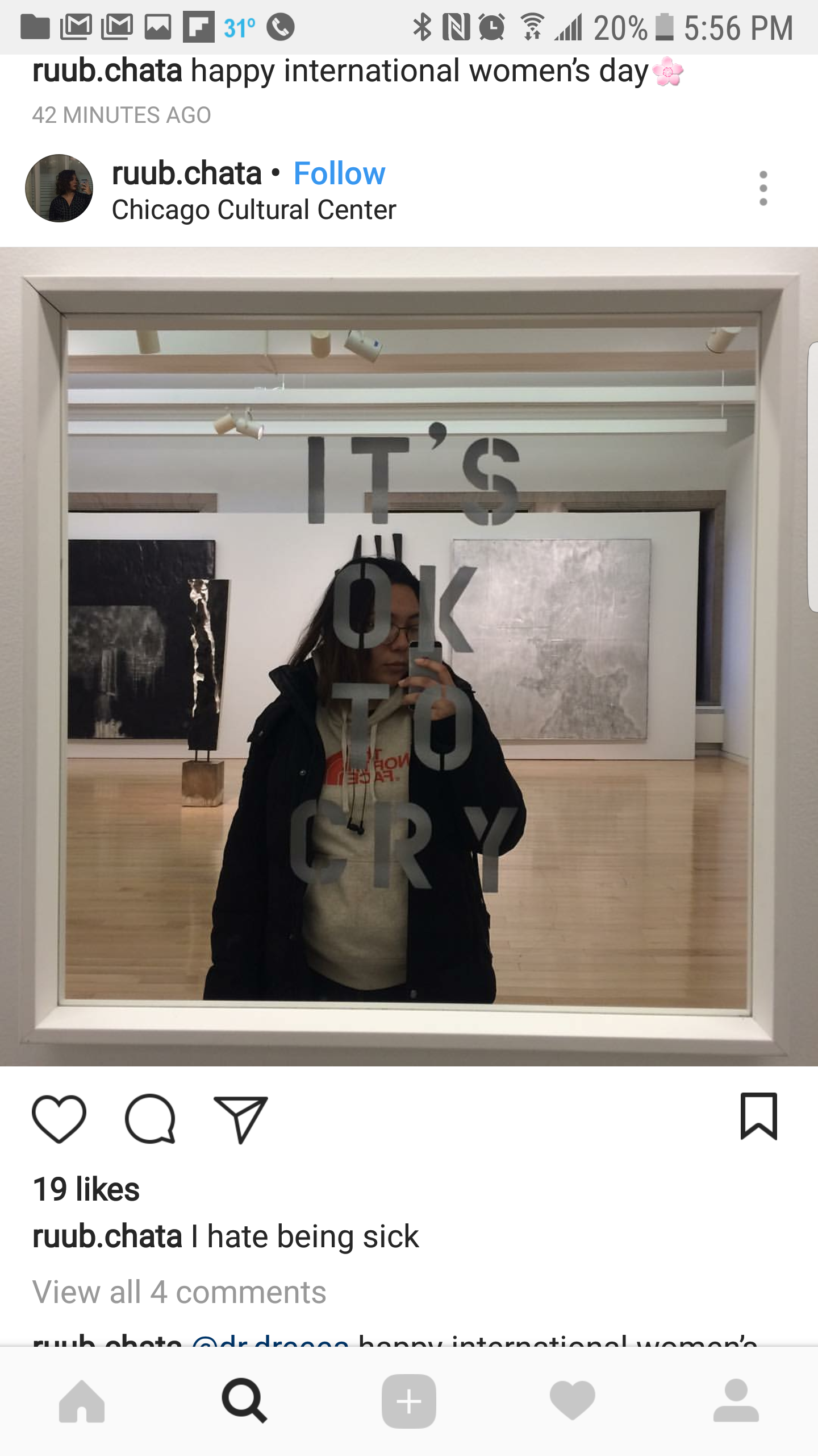 A person taking a selfie in a mirror at an art gallery. The mirror has the phrase 'IT'S OK TO CRY' written on it. The person is wearing glasses, a hoodie, and a jacket. The background shows an art exhibit with paintings and sculptures in a modern gal