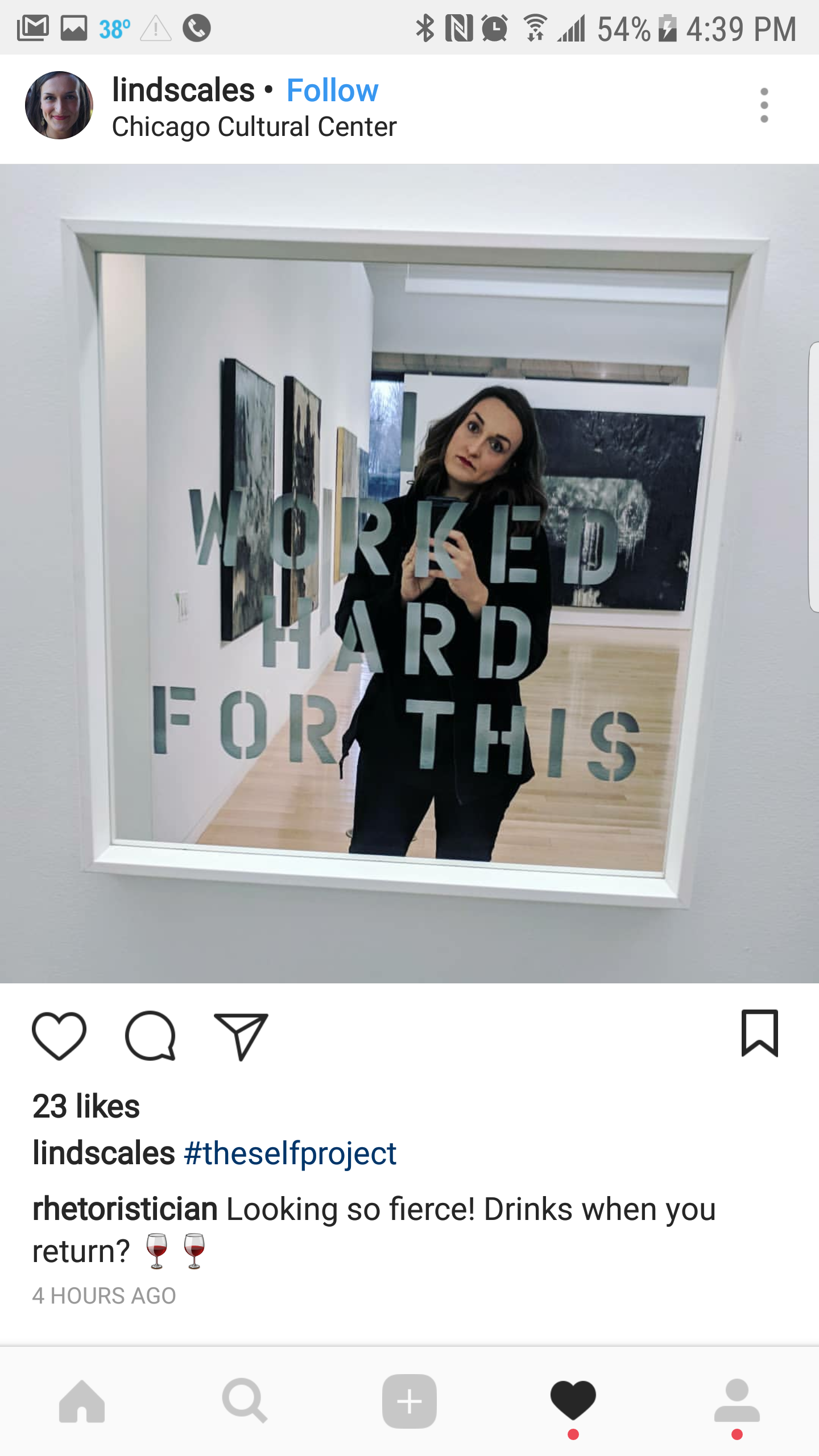 A woman taking a selfie in a mirror with the words "WORK HARD FOR THIS" on it. She is in an art gallery with paintings on the walls.