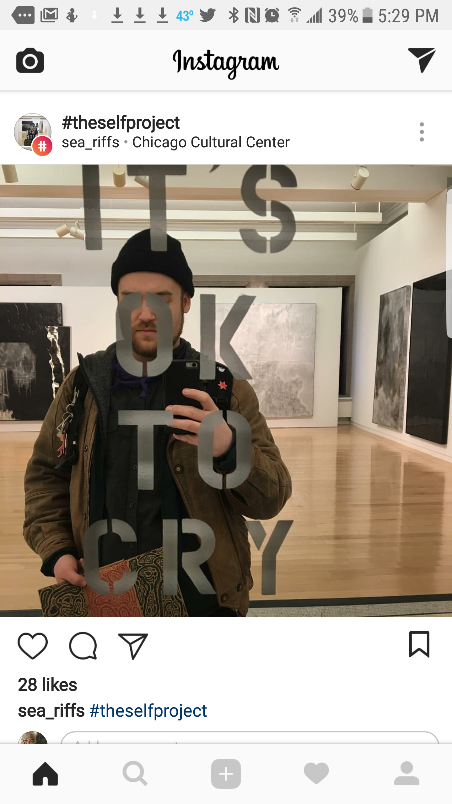 A man taking a mirror selfie in an art gallery with paintings on the wall behind him. He is wearing a black beanie and a brown jacket, holding a patterned item in his left hand. The mirror has the words 'IT'S OK TO CRY' written on it.