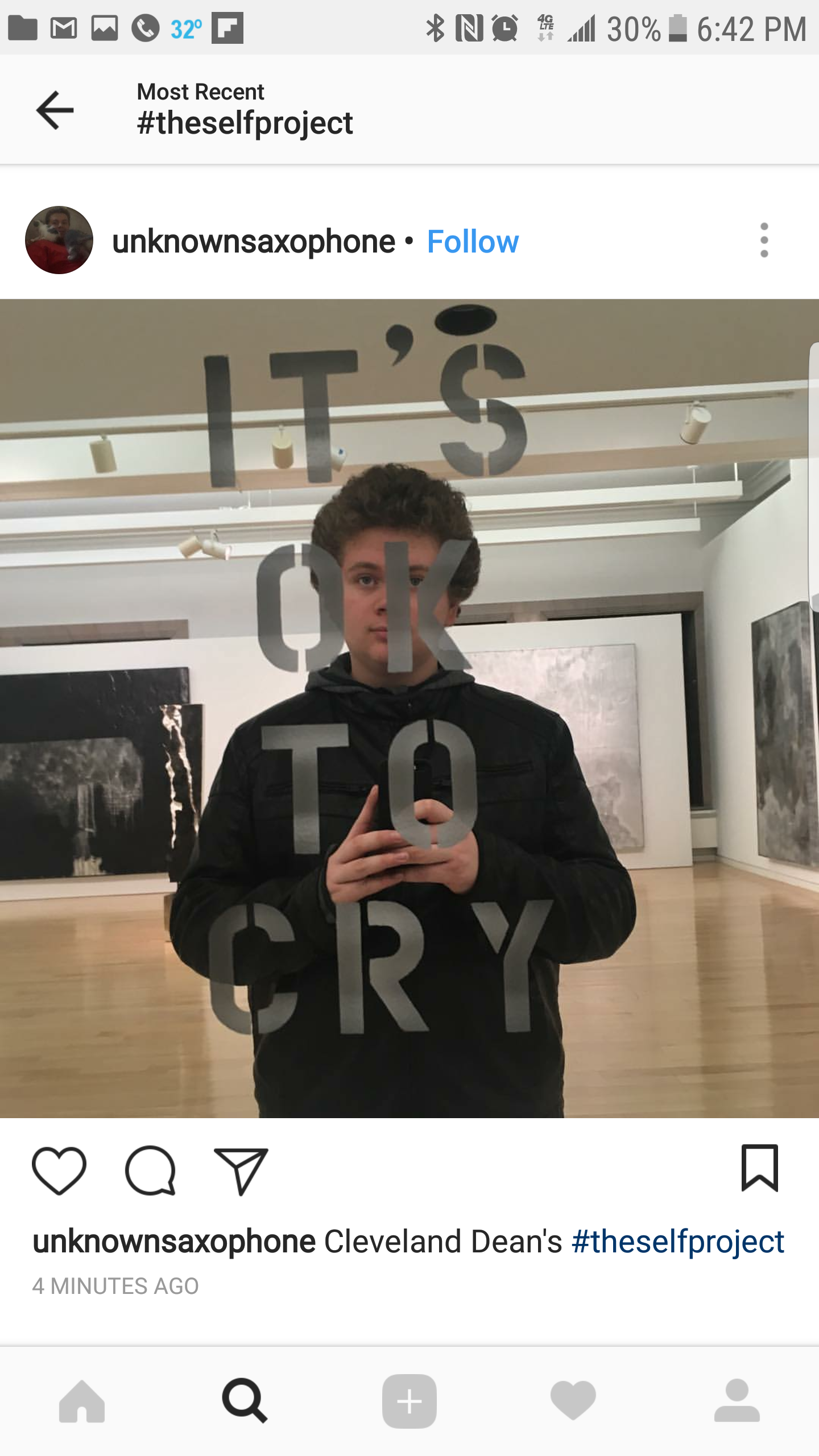 A person taking a mirror selfie in an art gallery, with the words 'IT'S OK TO CRY' written on the glass mirror.