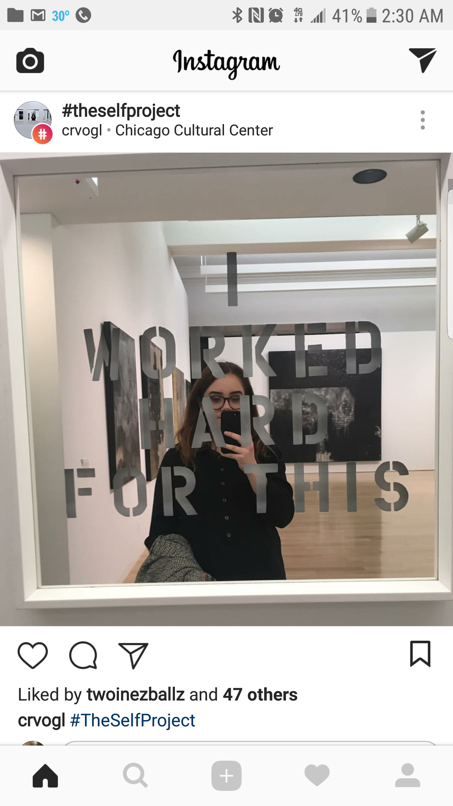 A woman taking a selfie in a mirror with the words 'I WORKED HARD FOR THIS' on it, in an art gallery.