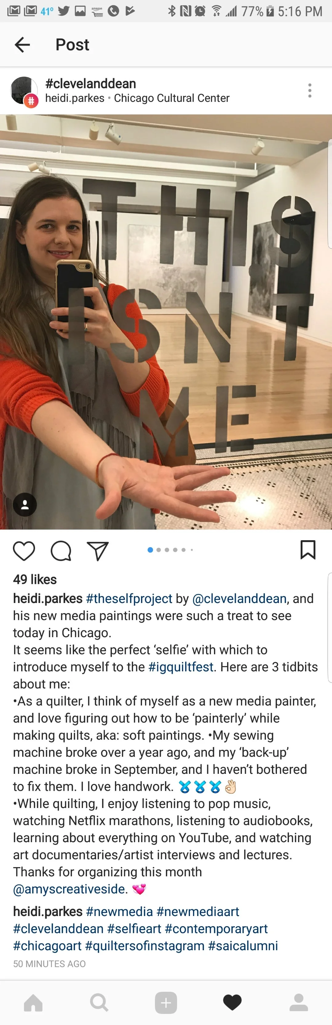 A woman taking a selfie in a gallery or art space with a mirror engraving that reads "THIS IS MY SELFIE." She is smiling, has long brown hair, and is wearing a light gray top with an orange sweater. The background features wooden floors and artwork o