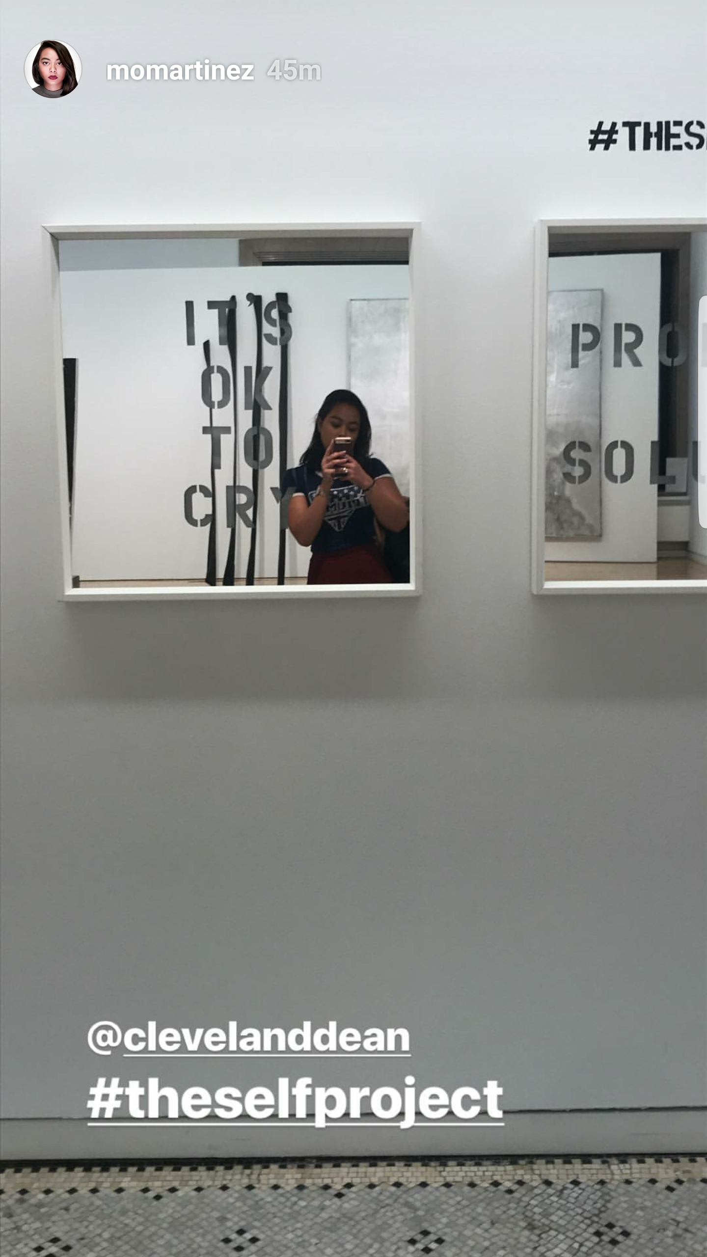 A woman taking a mirror selfie in an art gallery or museum with various artworks and text on the walls, reflecting in multiple mirrors.