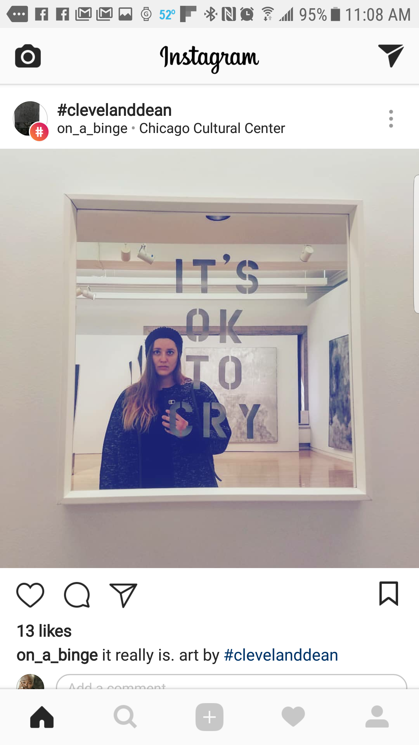 A woman taking a selfie in a museum mirror with the text "IT'S OK TO CRY" on the glass. Art is visible in the background.