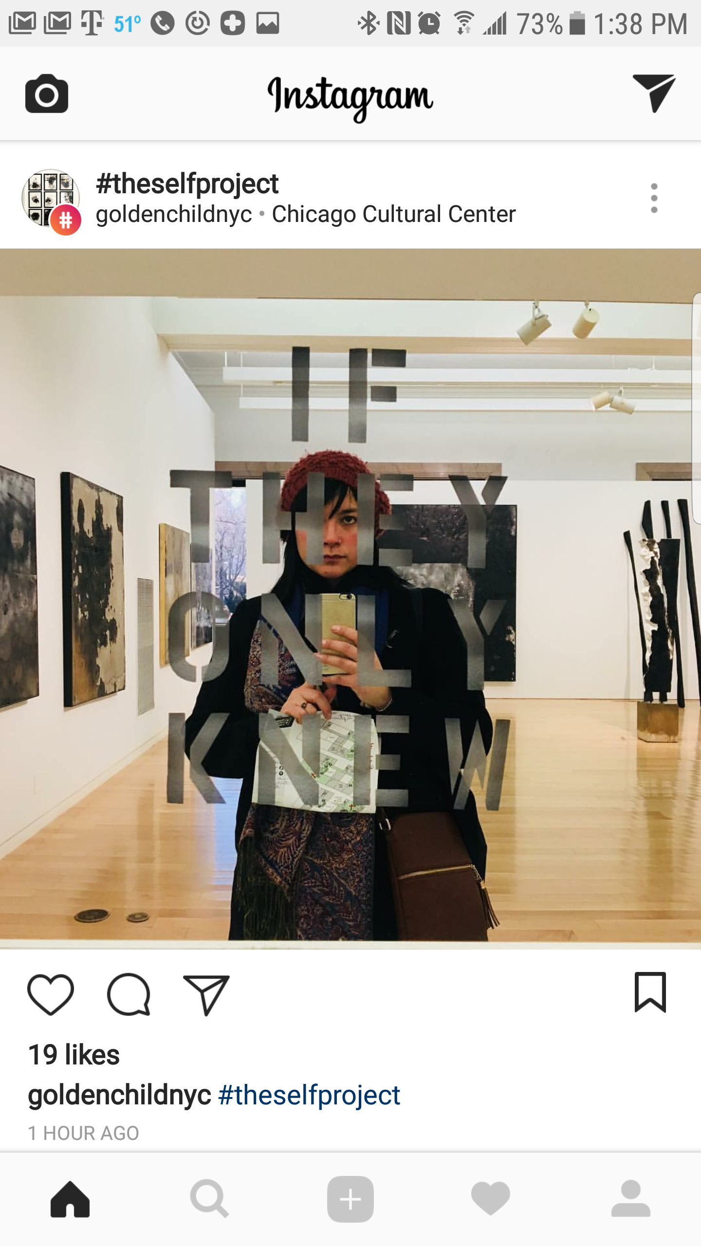 A person taking a selfie in an art gallery using a mirror that has the words 'THE SELF PROJECT' written on it, with artwork displayed on the gallery walls behind them.