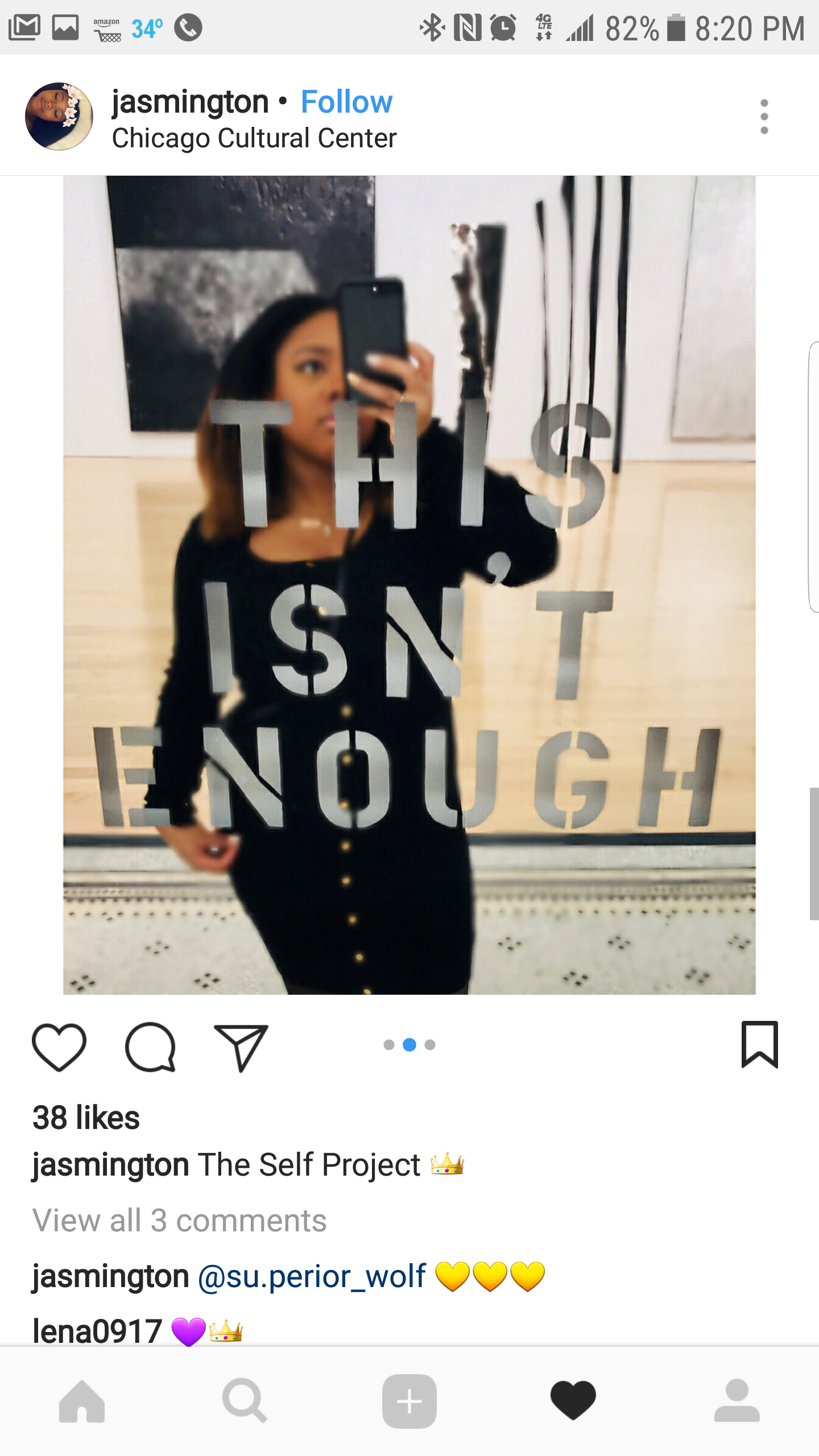 A woman takes a mirror selfie in a mirror with the phrase 'This isn't enough' written on it. She is wearing a black dress with gold buttons.