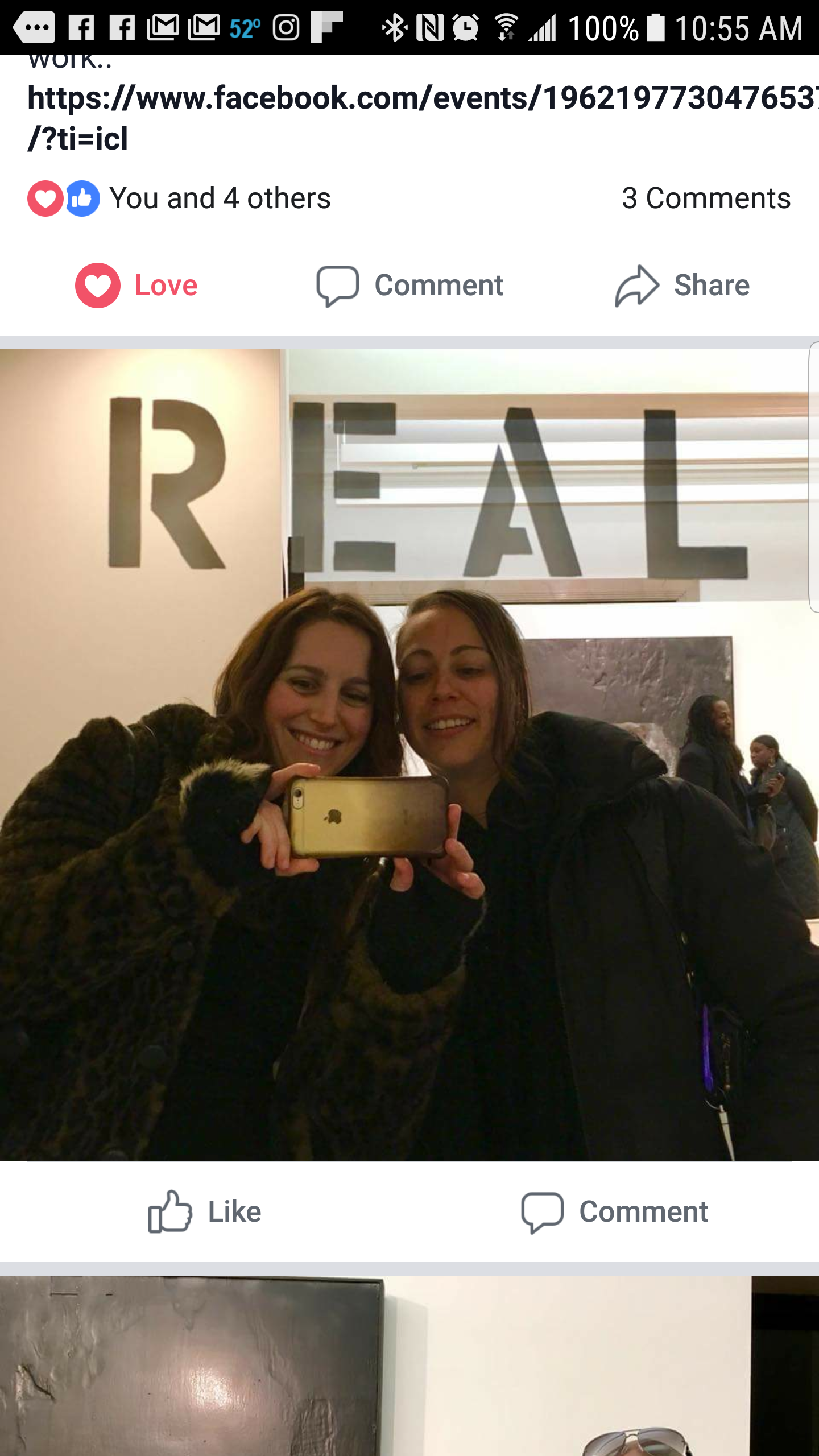 Two women taking a selfie in front of a mirror with large letters spelling 'REAL' on the wall behind them. Both women are smiling; one is wearing a leopard print coat and the other a black jacket.