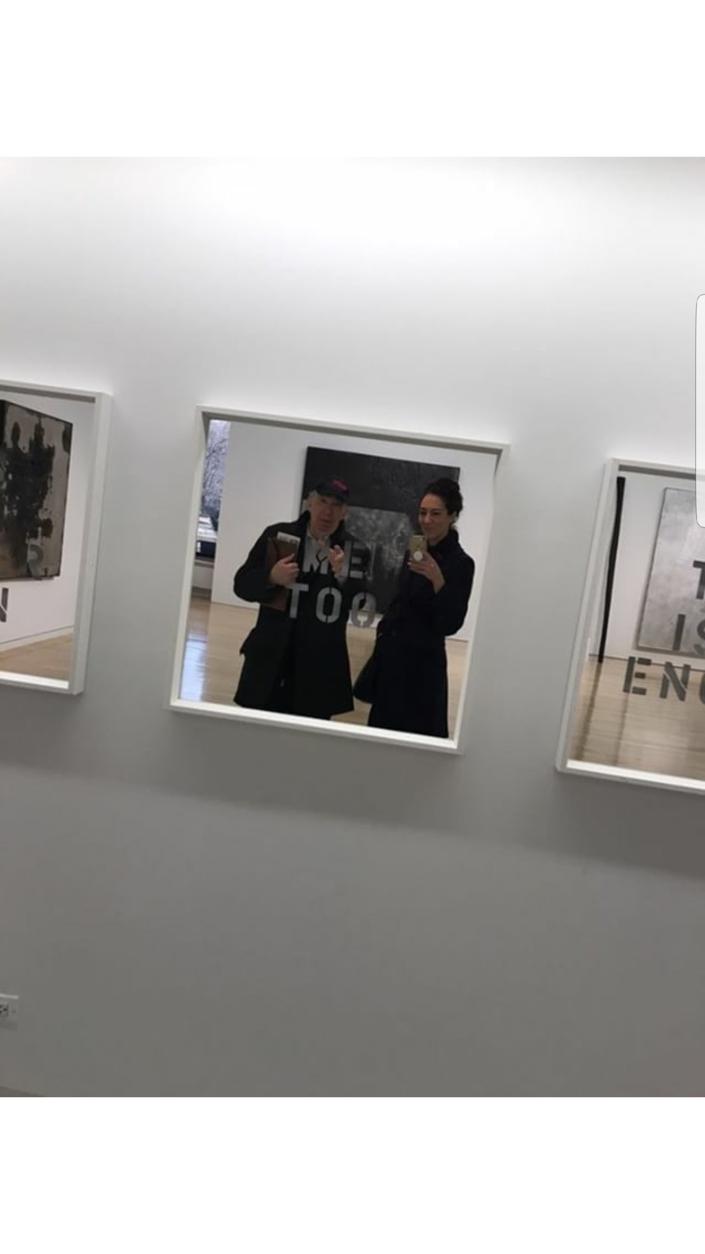 Two people are taking a mirror selfie in an art gallery, with framed artwork on the wall behind them.