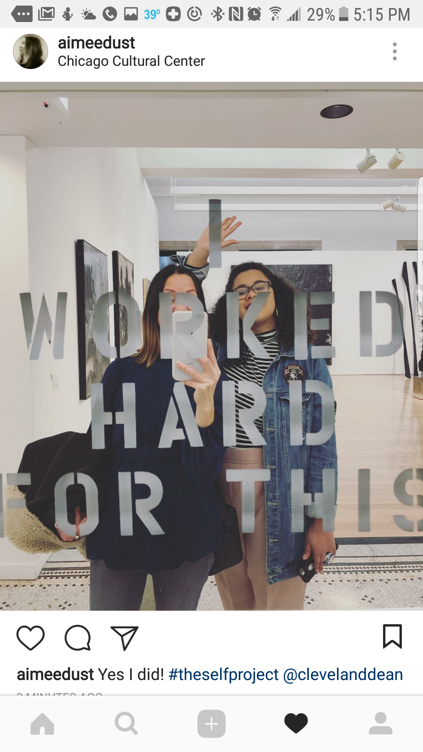 Two women taking a selfie in front of a mirror with the words "WORK HARD" and "OR" visible. The background is a gallery or art space at the Chicago Cultural Center.