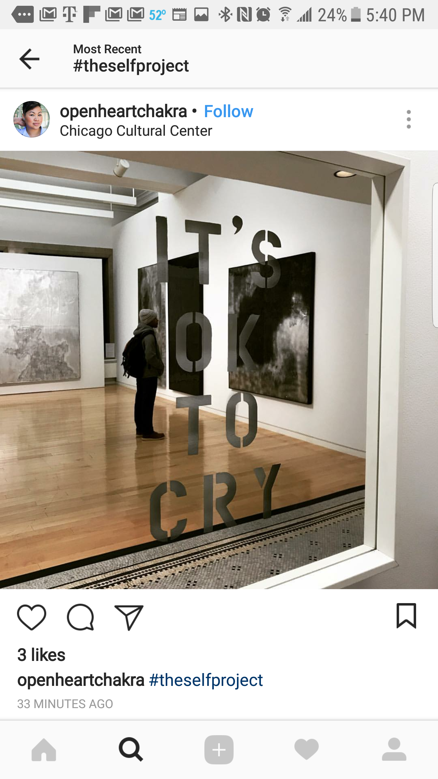 A person wearing a gray hat and backpack viewing artwork in a gallery, seen through a glass door with the text "IT'S OK TO CRY" written on it.