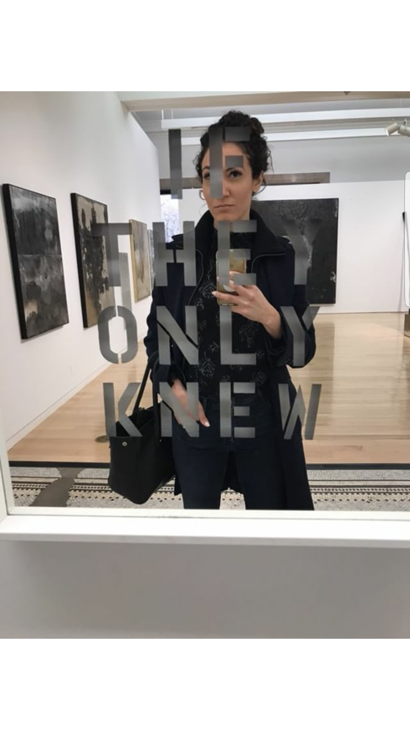 A woman taking a selfie in an art gallery, reflected in a mirror with the words 'THE ONLY KNEE' written on it.
