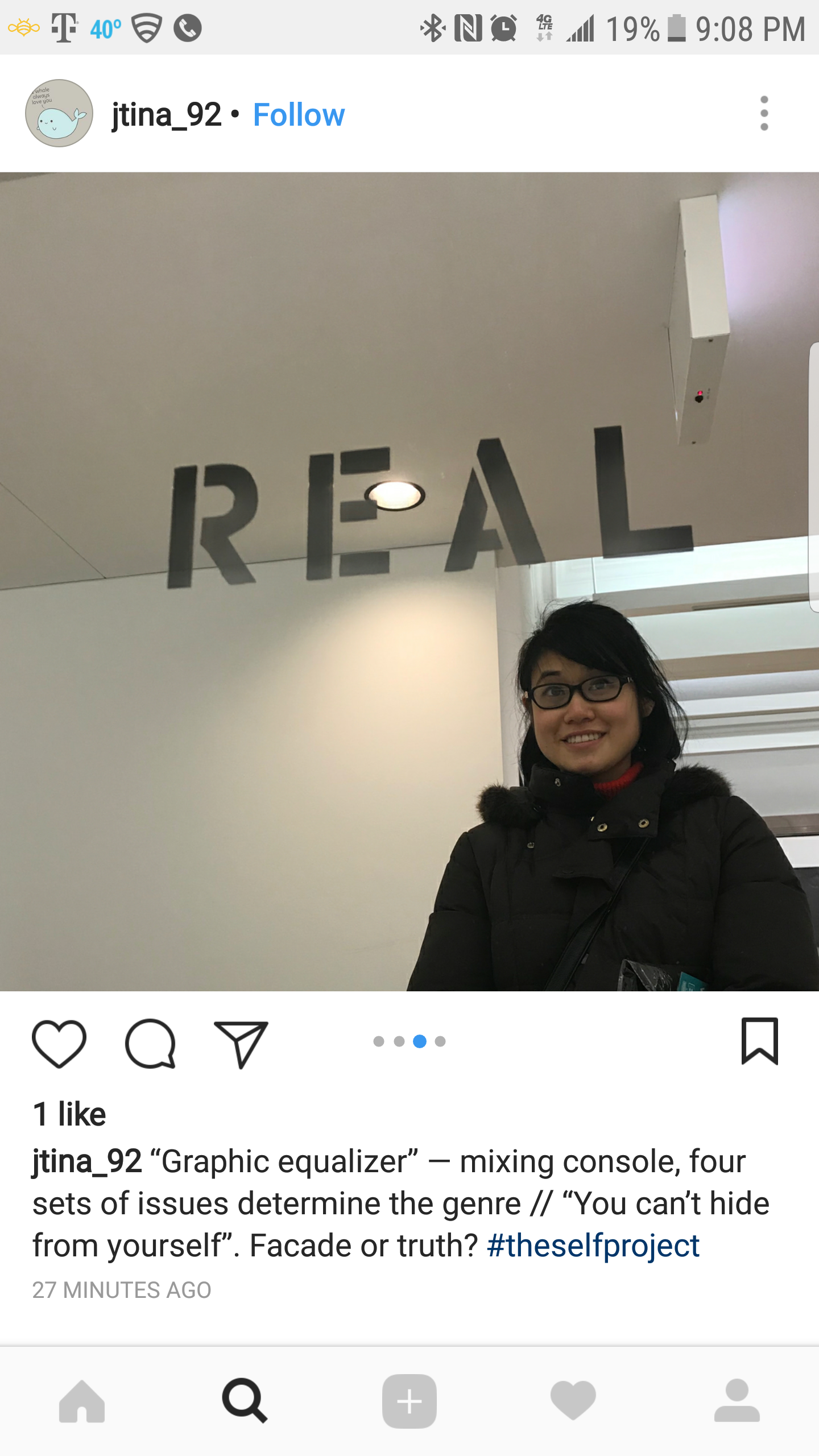 A woman wearing glasses and a black coat standing inside a building near a mirror with letters spelling 'REAL' on it. The woman is smiling and the ceiling has a light fixture.