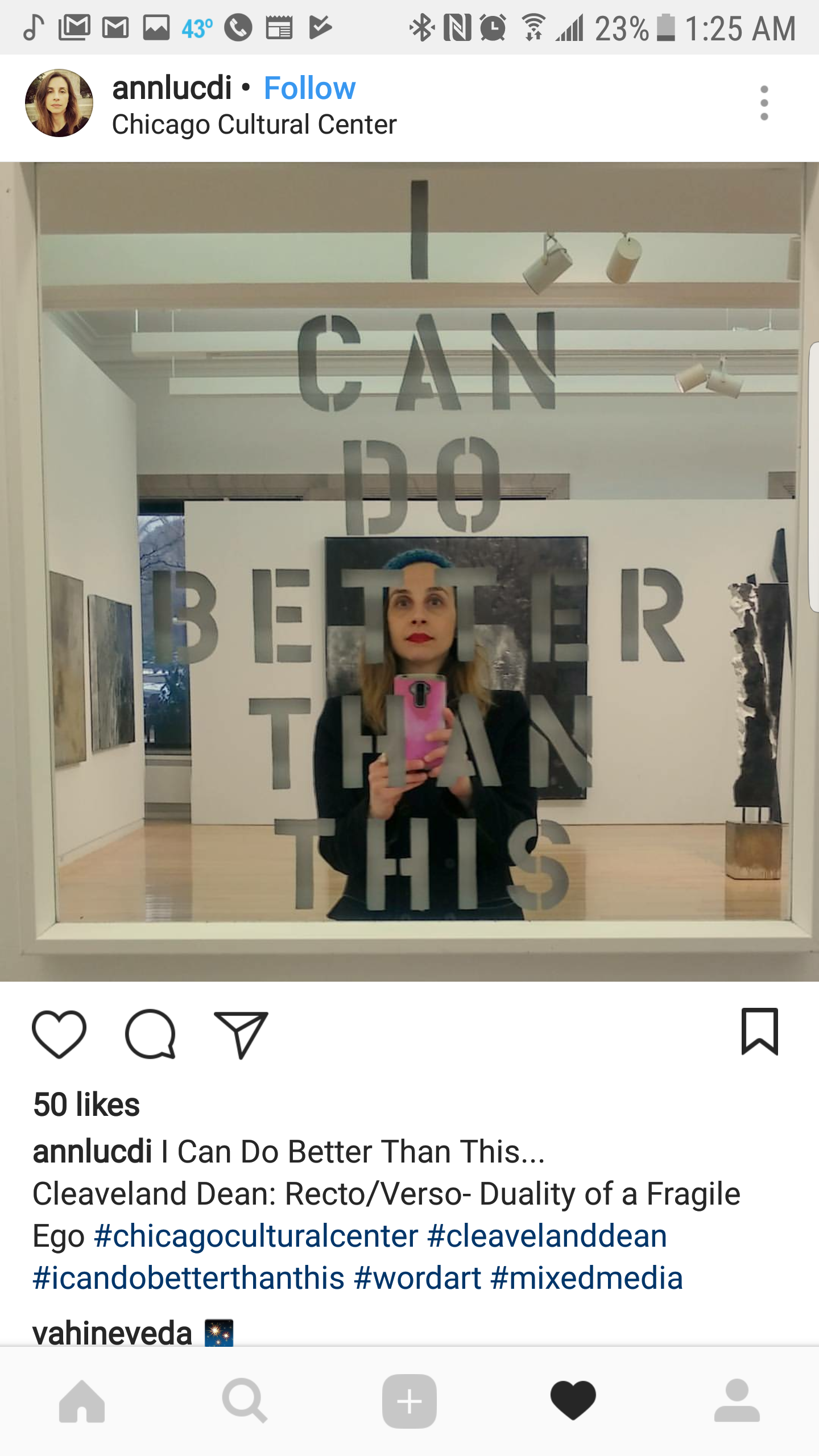 A woman taking a mirror selfie with a pink camera in an art gallery. The mirror has a quote on it that reads, "I CAN DO BETTER THAN THIS." Several artworks are visible in the background within the gallery.