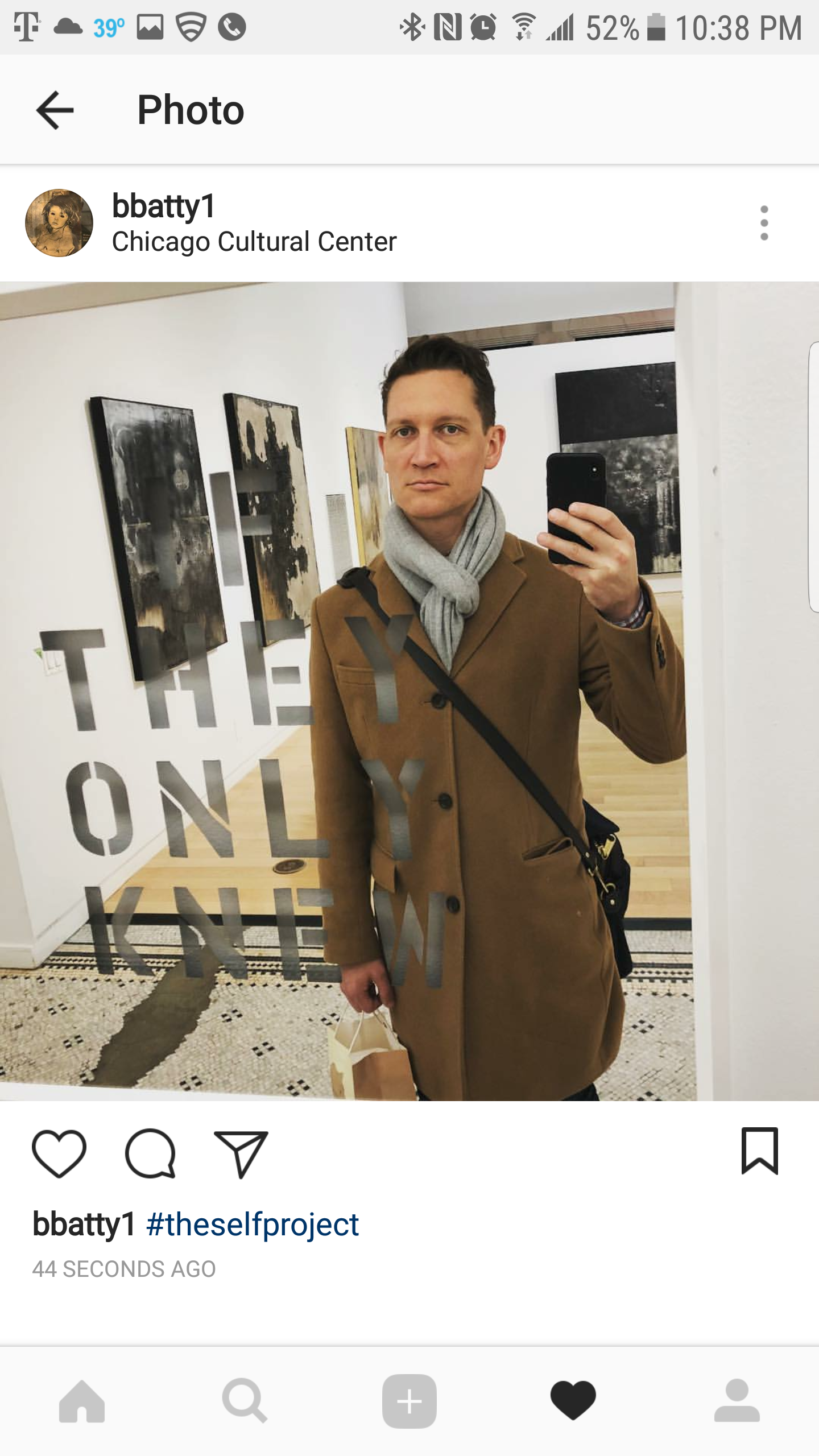 A man taking a mirror selfie at the Chicago Cultural Center, with black and white artwork on the walls behind him, wearing a brown coat and gray scarf.